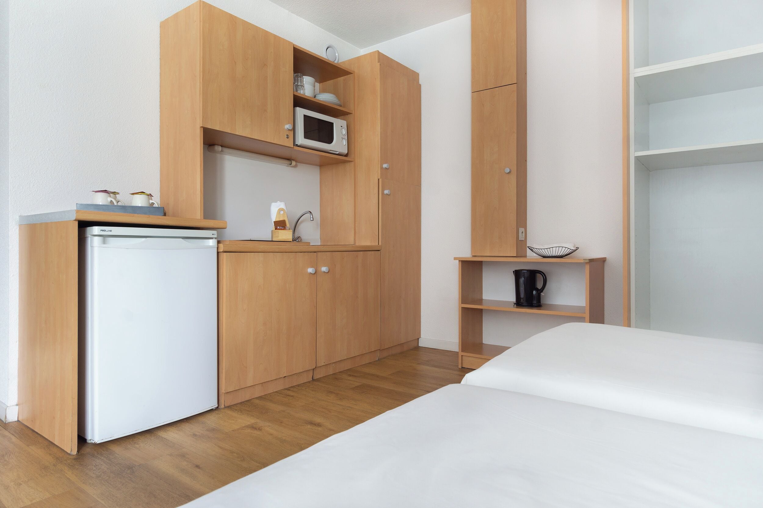 A compact room with a bed, a kitchenette with a microwave, a small coffee maker, and a shelf with a kettle.