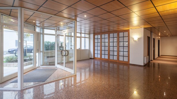 Un hall d’entrée spacieux avec un sol poli, des portes vitrées donnant sur un espace extérieur et un plafond carrelé.