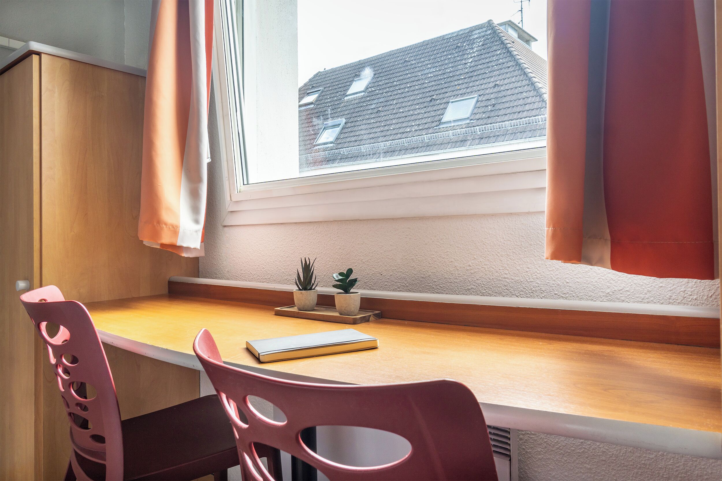 A room with a wooden desk, two chairs, a window with curtains, and two potted plants on the windowsill.