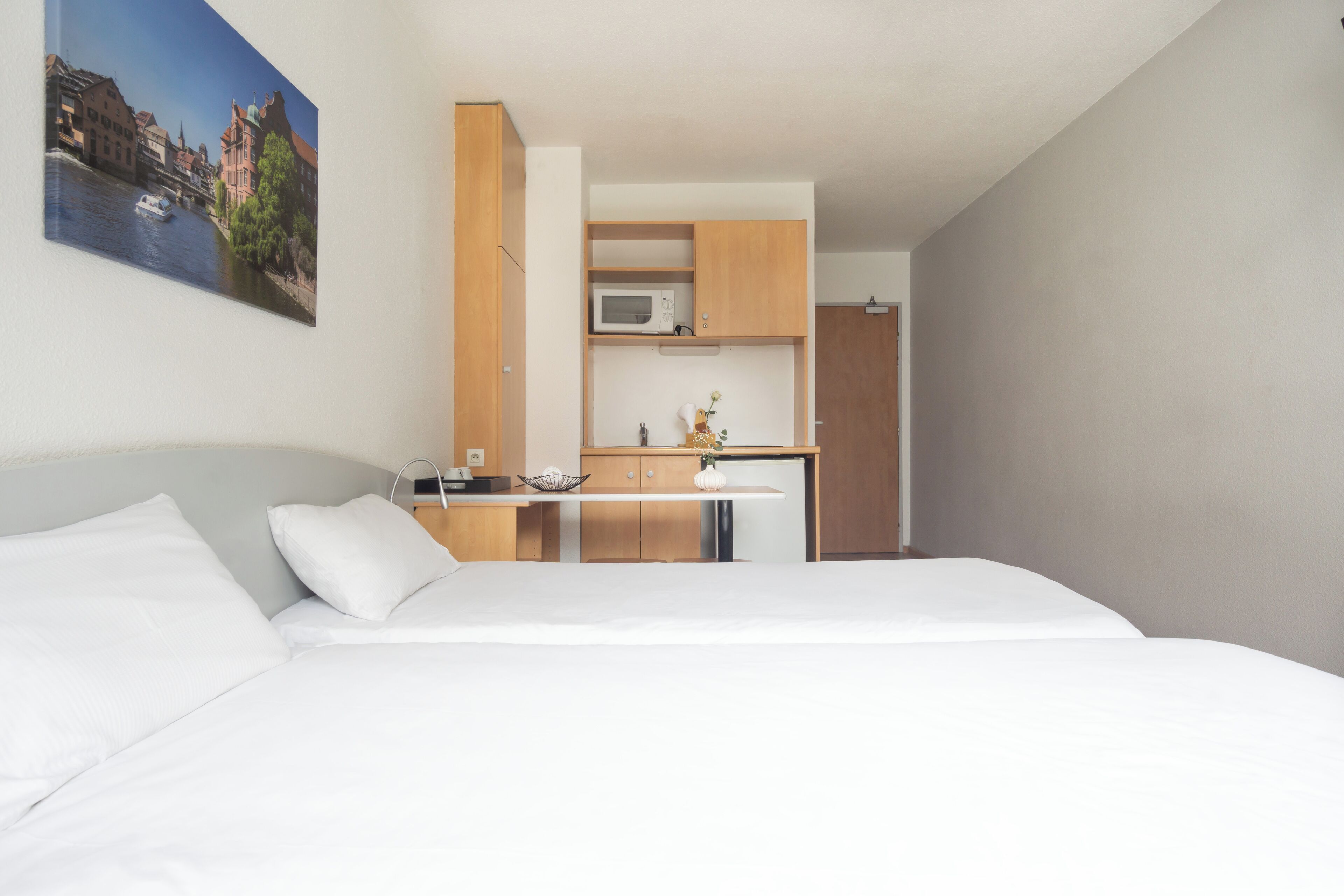 A modern hotel room with a large bed, a small kitchenette, and a painting of a river scene on the wall.