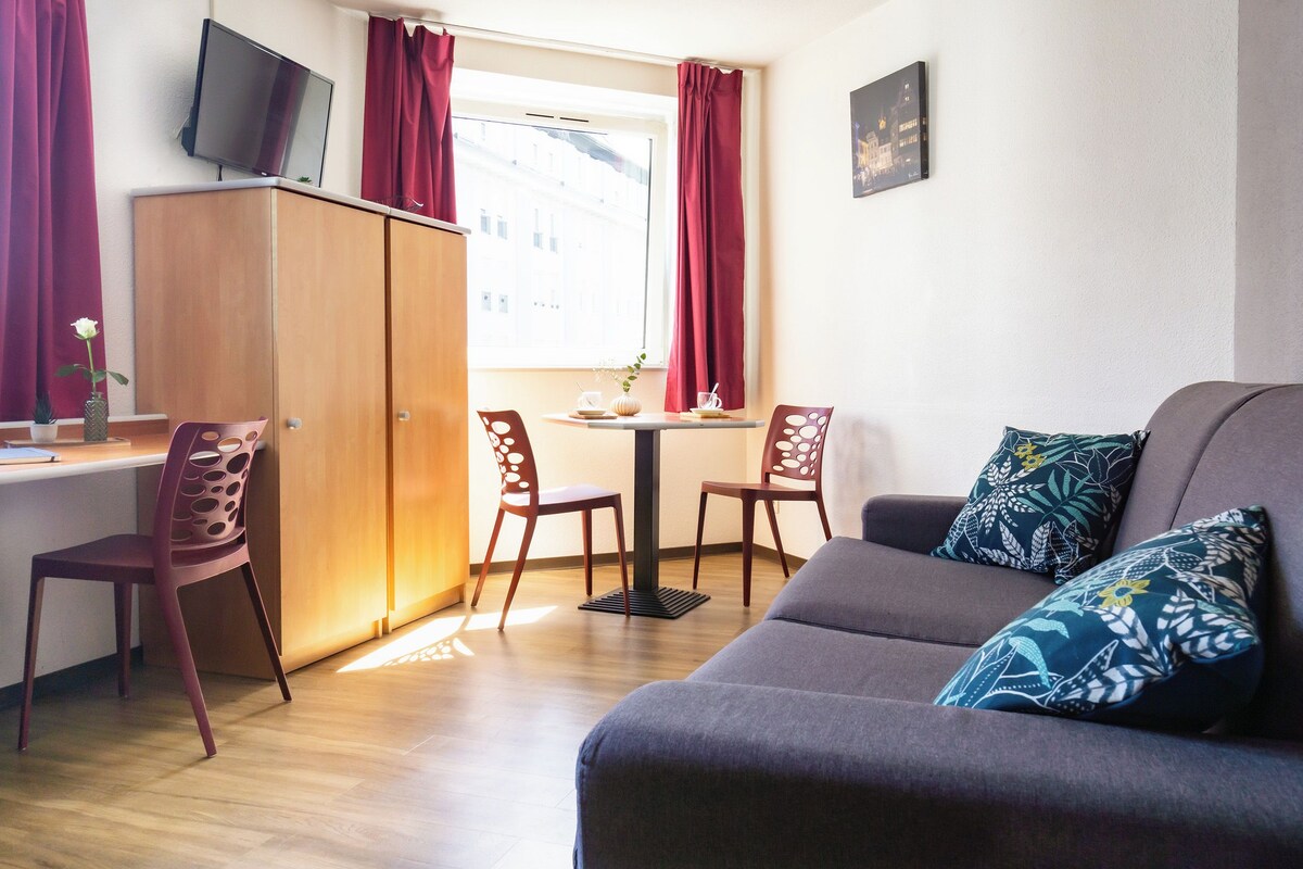 A room with a sofa, two chairs, a small table, a TV on the wall, and a window with curtains.