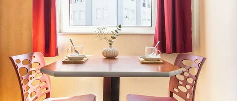 A dining area with a table, two chairs, a vase with flowers, and a window with curtains.