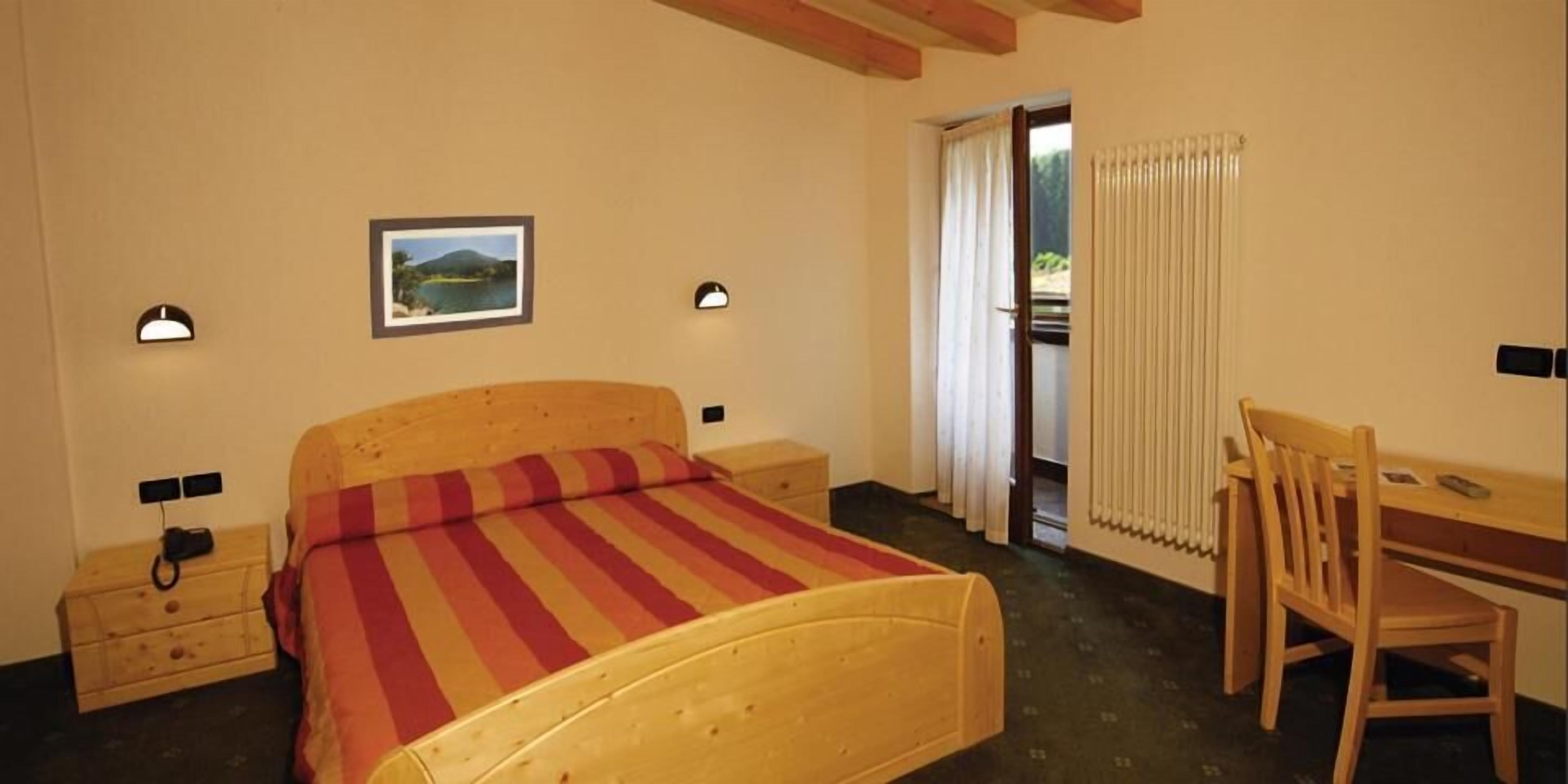 A bedroom with a wooden bed, a desk, and a chair. There is a window with curtains, a radiator, and a picture on the wall.