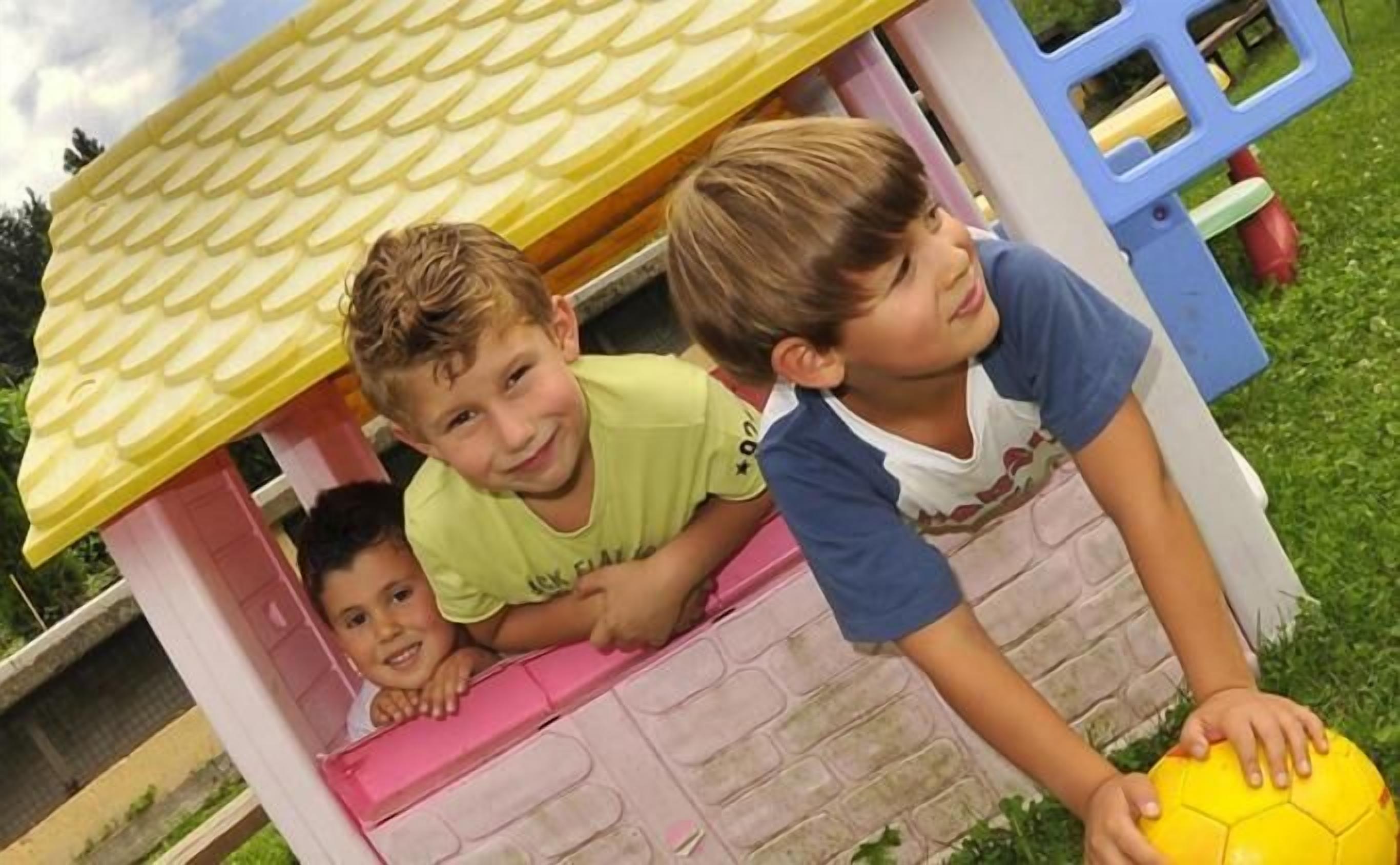 Tres niños jugando afuera de una casita de juegos pequeña, rosa y amarilla con techo amarillo.