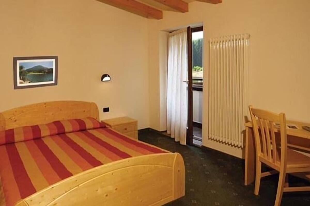 A bedroom with a wooden bed, a small bedside table, a chair, a radiator, and a window with curtains.