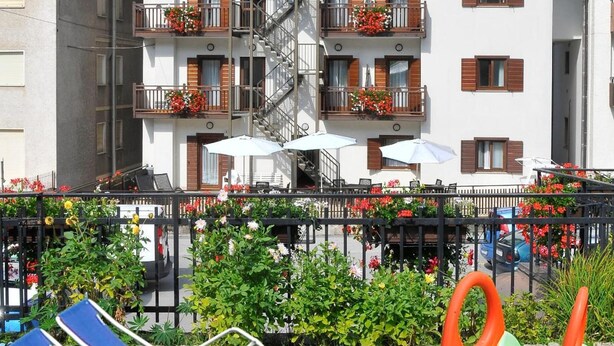 Un edificio de varios pisos con balcones, un jardín vallado con tumbonas y juegos infantiles, y un cielo despejado.