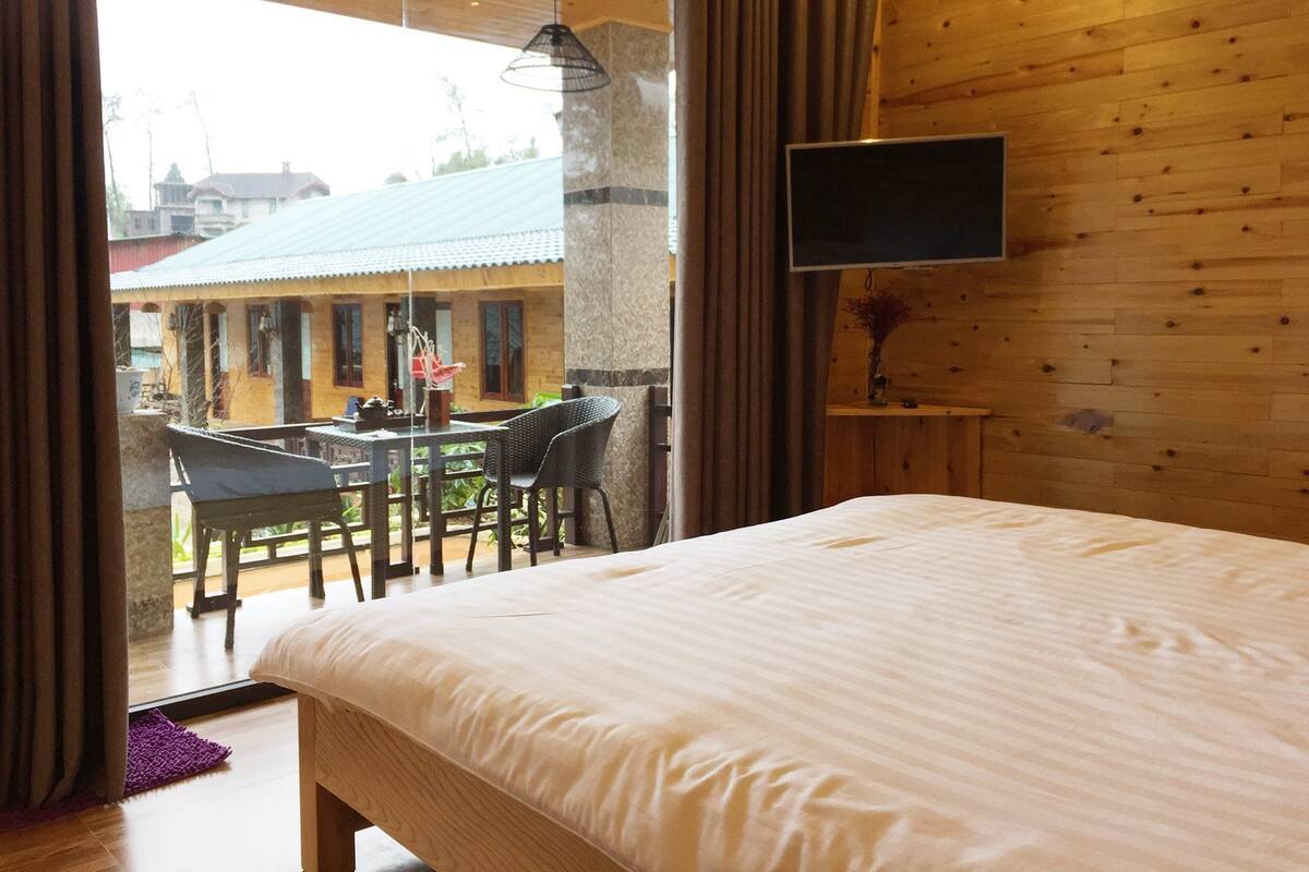 A bedroom with a large bed, a TV mounted on the wall, and a balcony with a table and chairs.