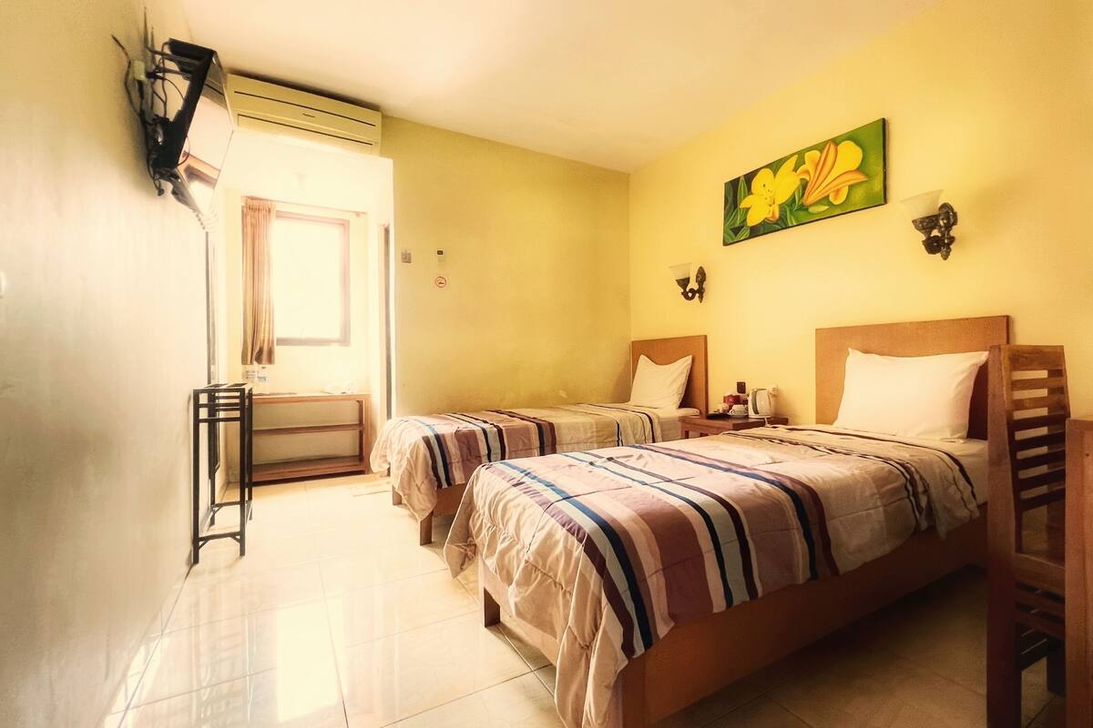 A hotel room with two beds, a desk, and a chair. There is a painting on the wall and a window with curtains.