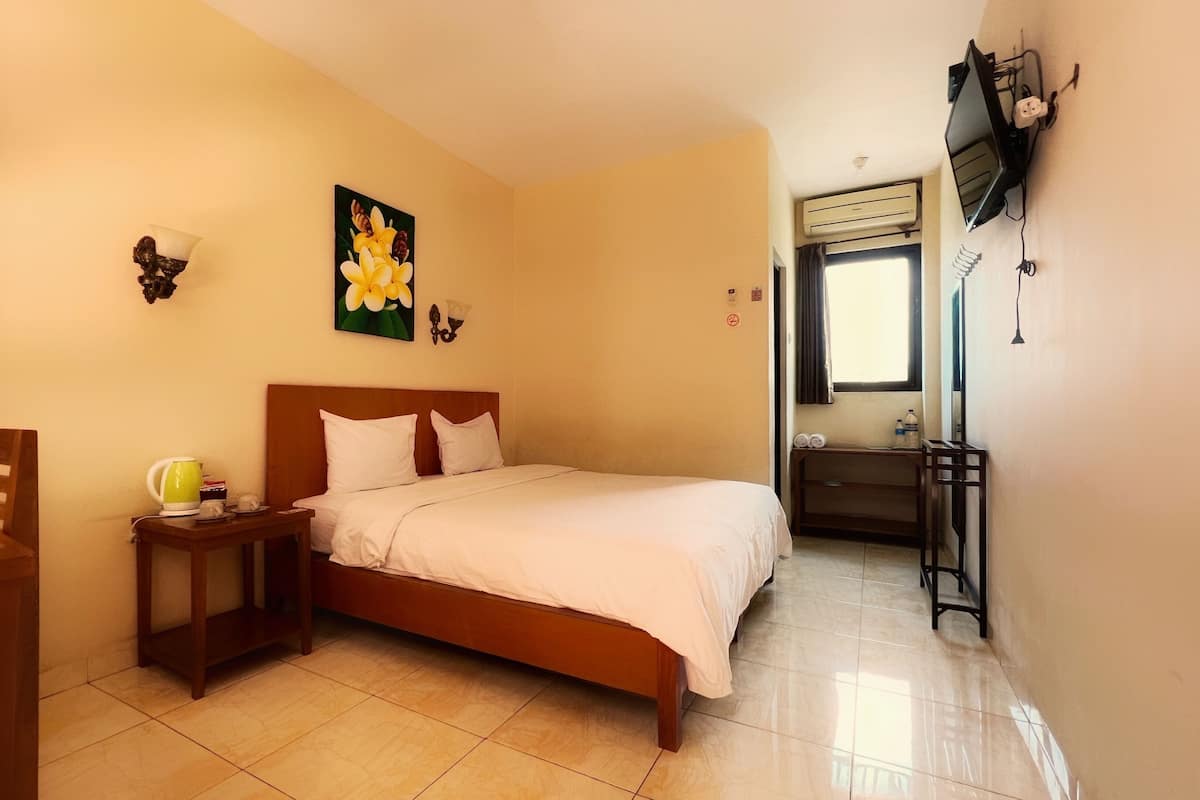 A hotel room with a bed, a small table with a kettle, a chair, a painting on the wall, and a window with curtains.