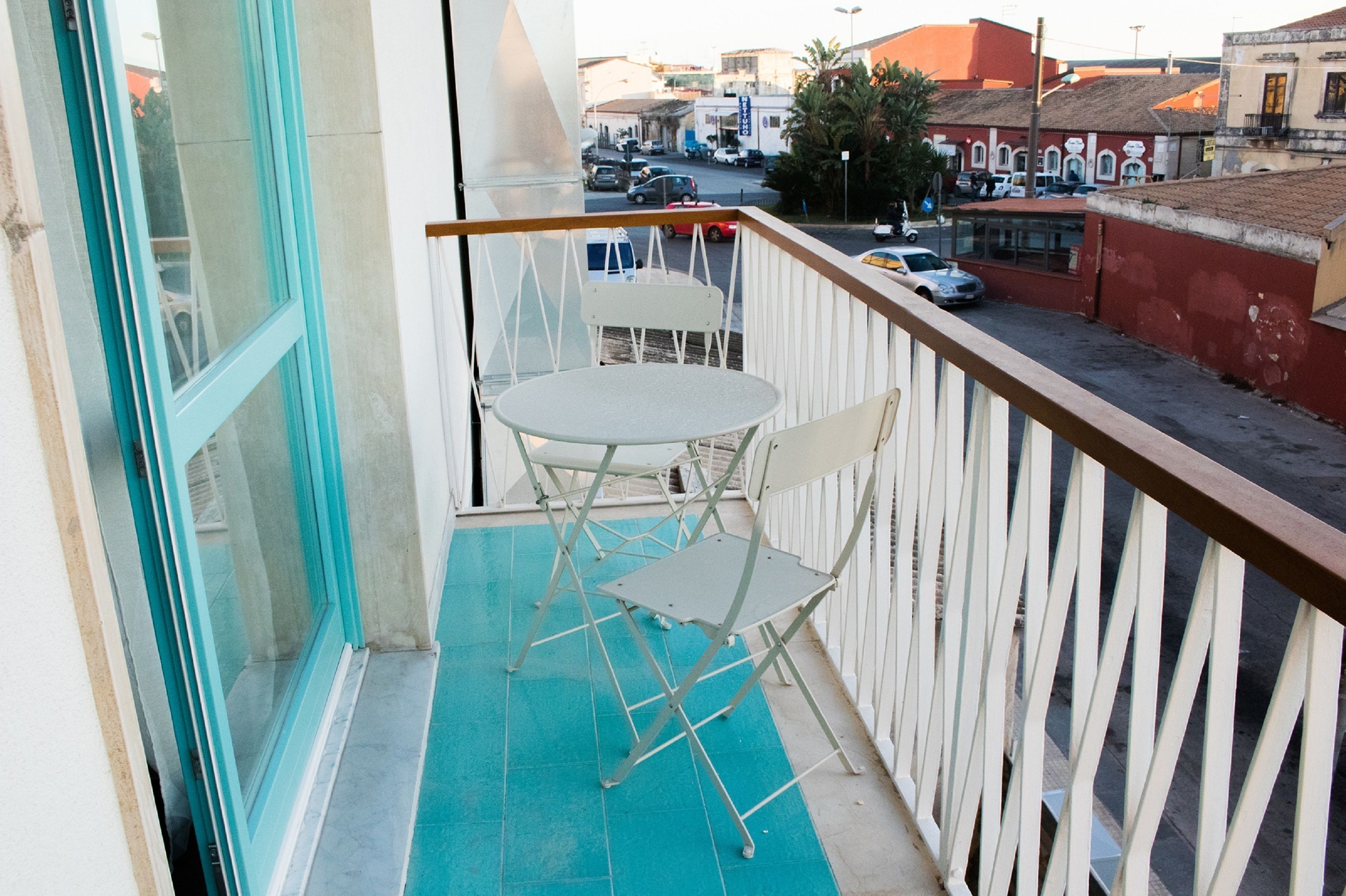 A balcony with a table and two chairs, overlooking a street with parked cars and buildings.