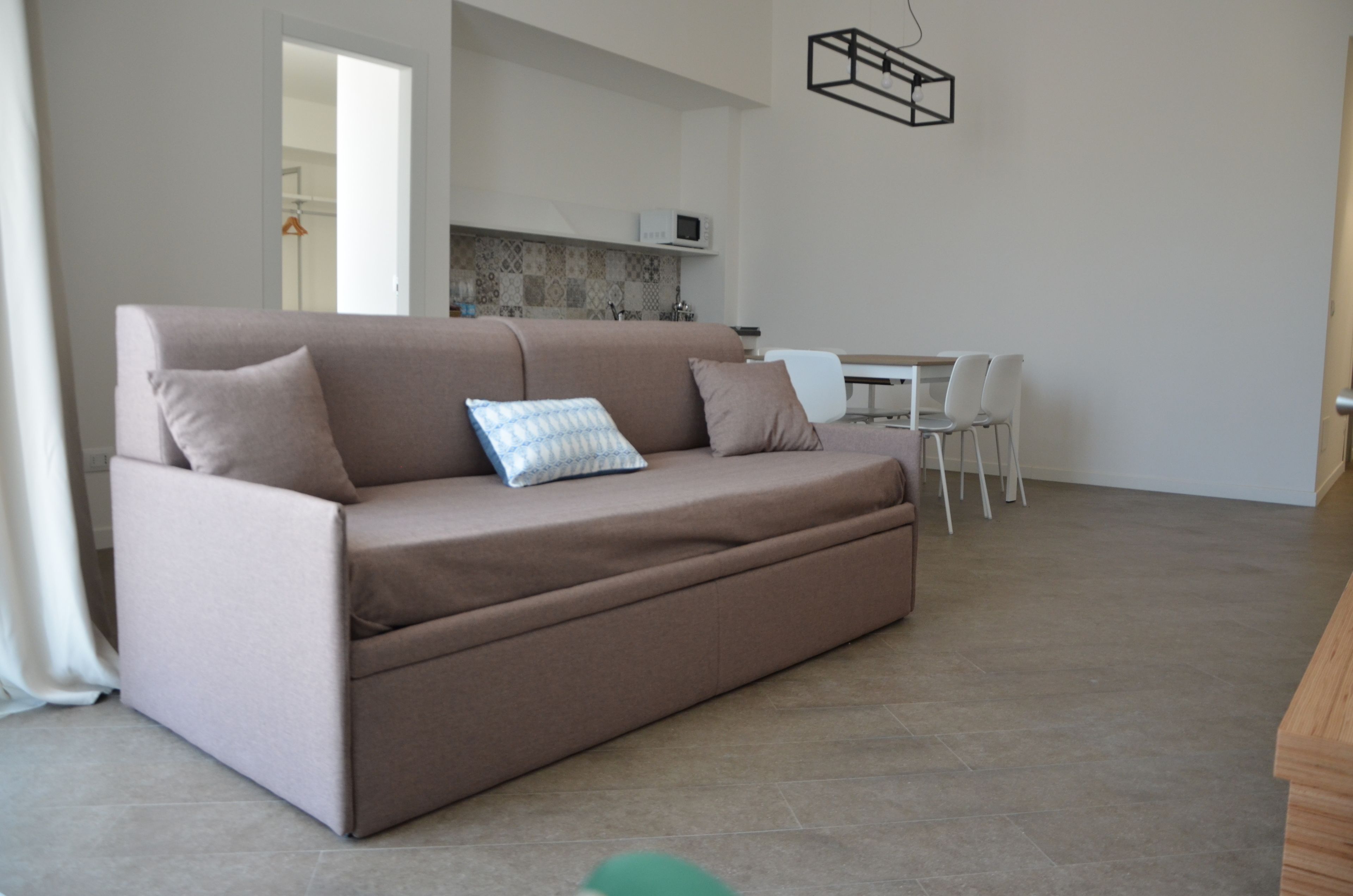 A modern living room with a sofa, a dining table with chairs, a kitchen area, and a hanging light fixture.