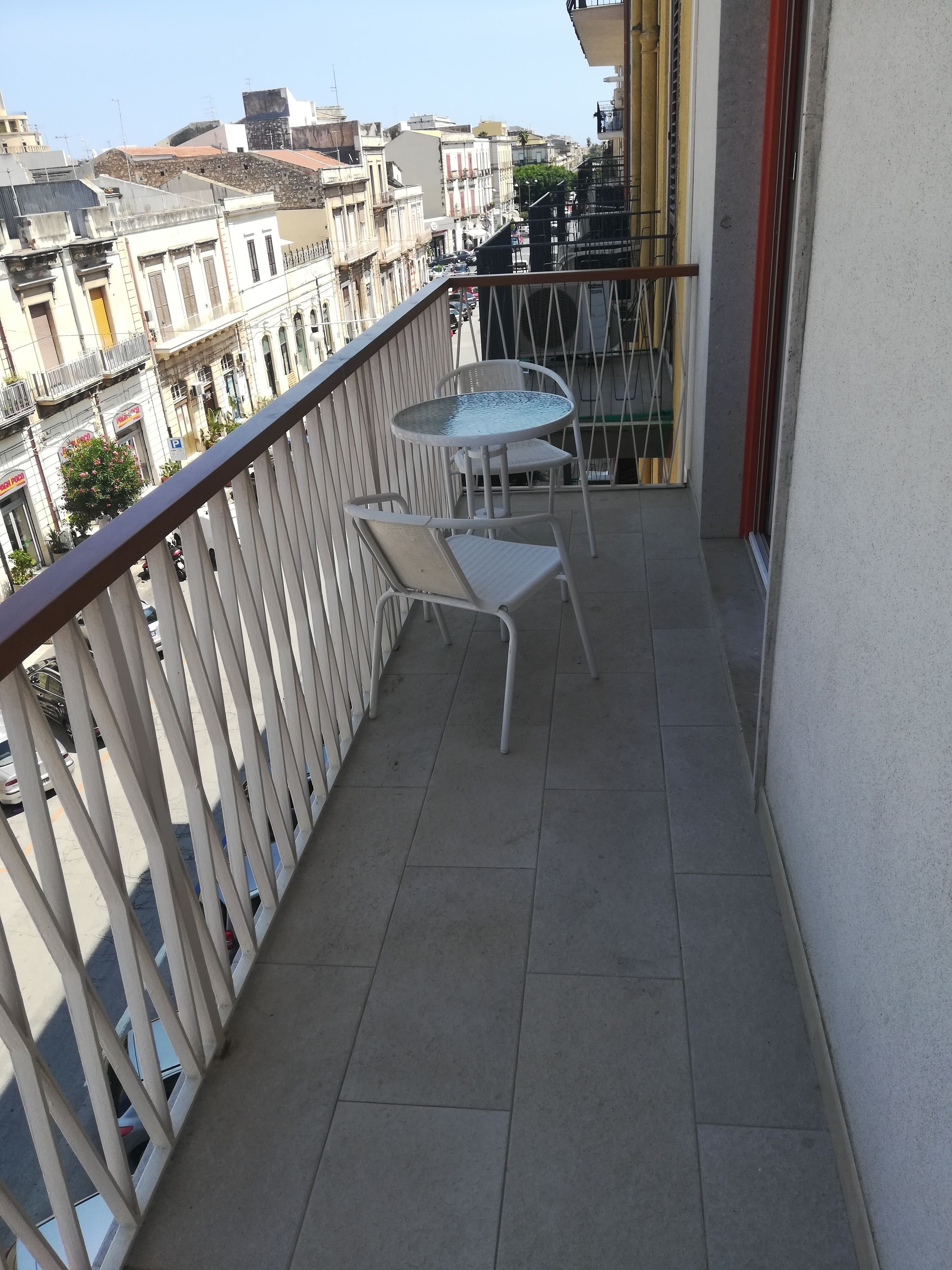 A balcony with a glass table and two chairs, overlooking a cityscape with buildings and parked cars.