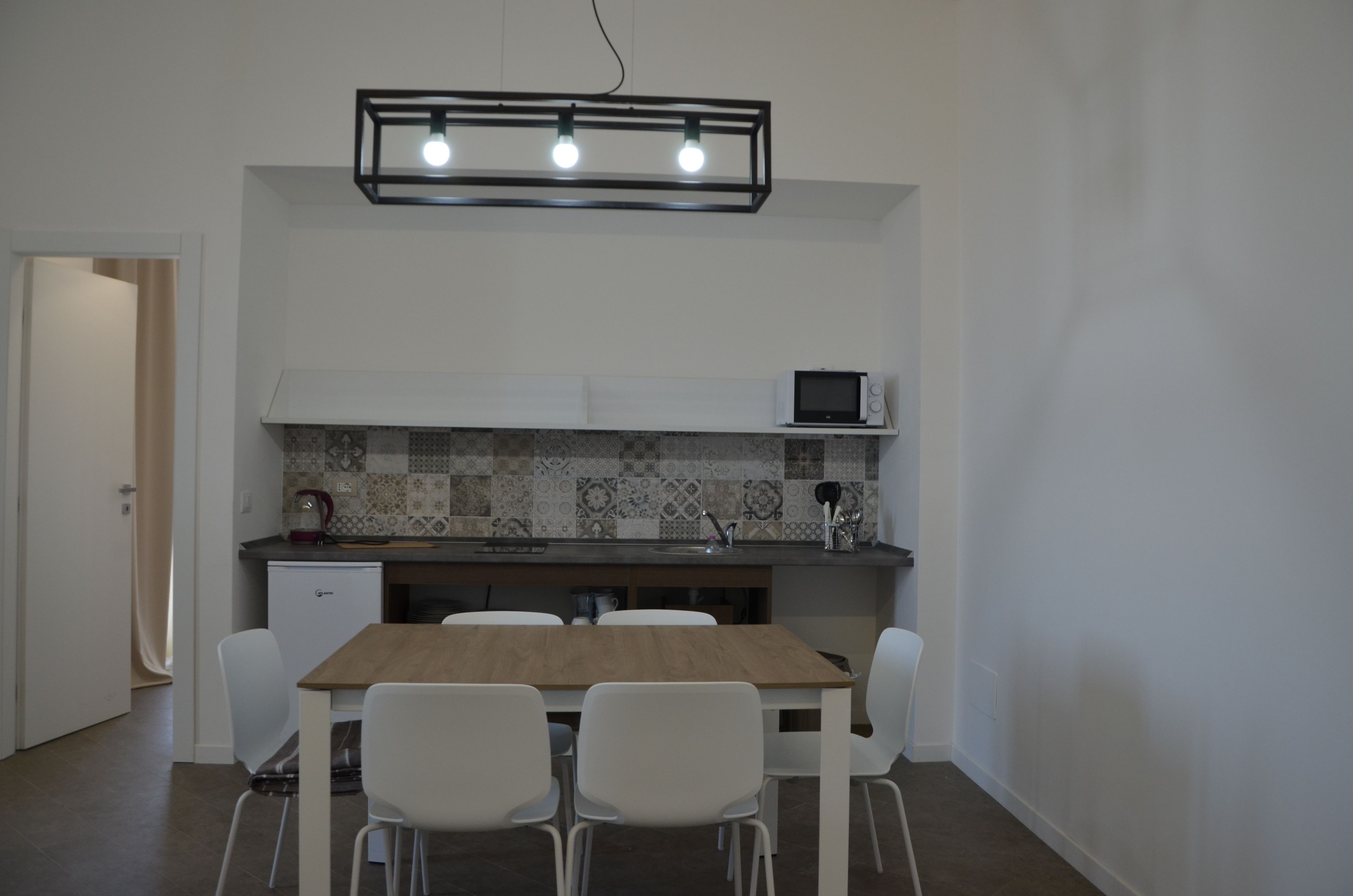 A compact kitchen with a microwave, a small refrigerator, a tiled backsplash, and a wooden dining table with four chairs.
