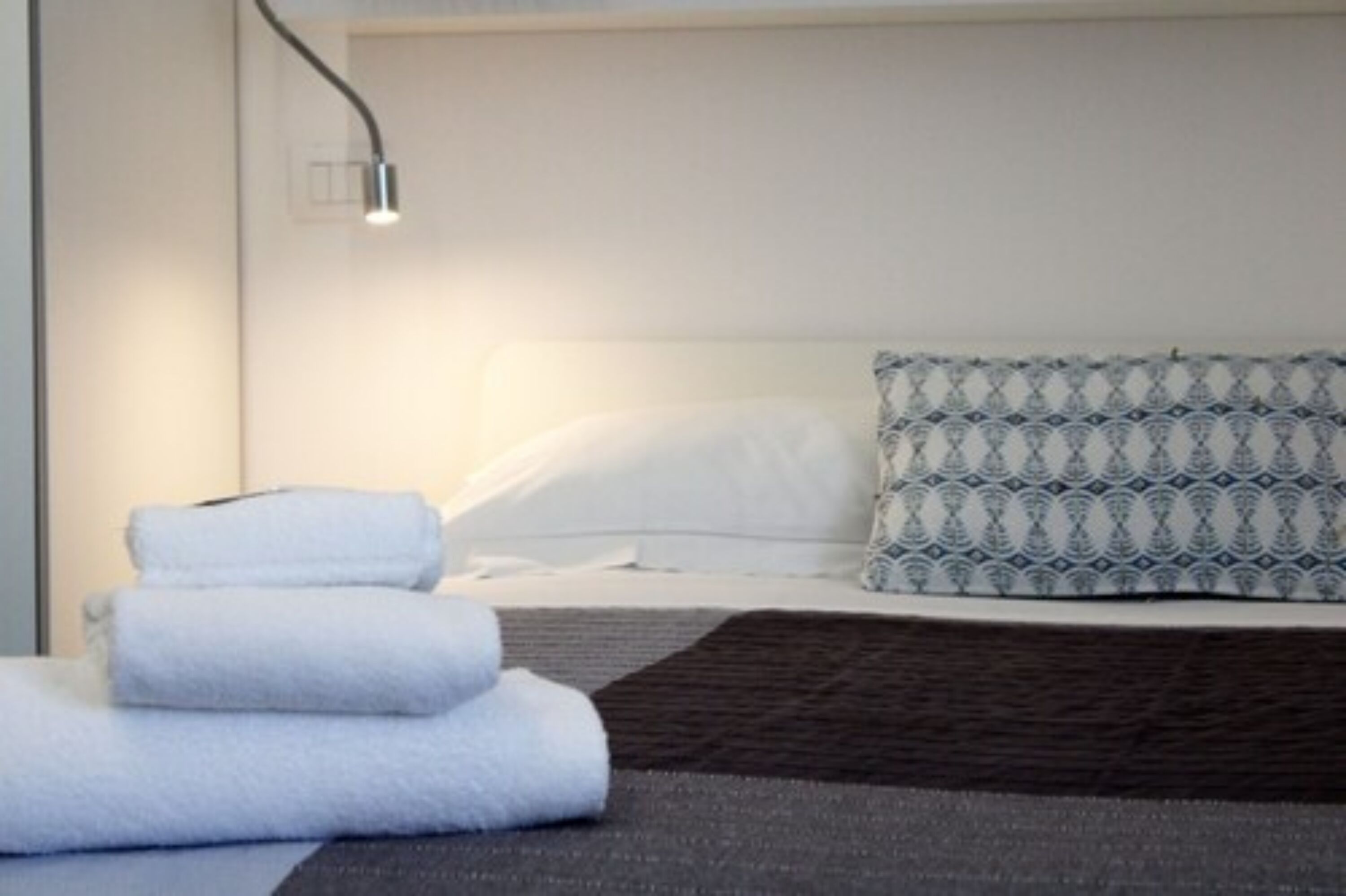 Una cama bien hecha con almohadas blancas y estampadas, una manta marrón oscuro y una pila de toallas blancas en el suelo.