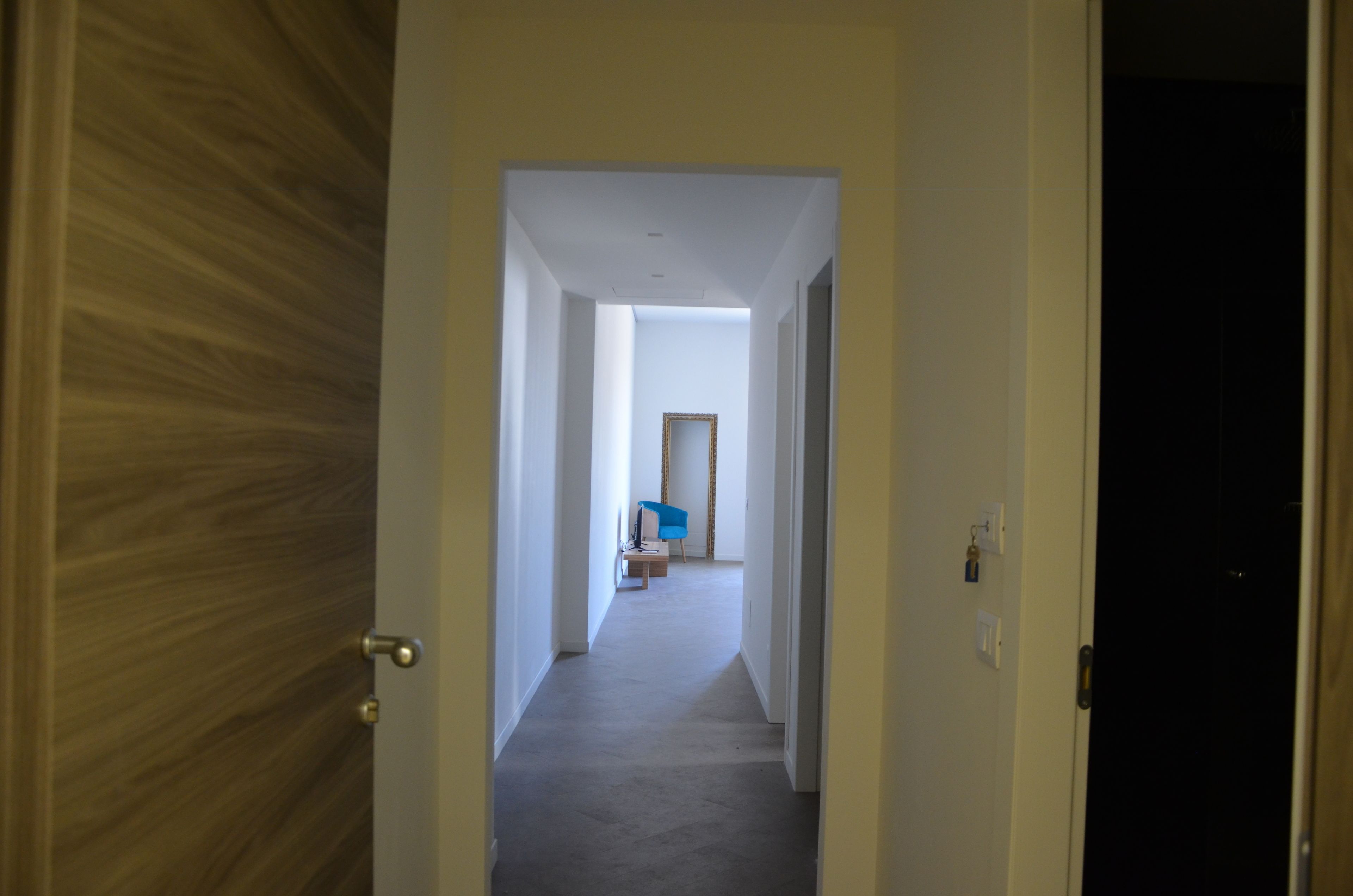 Un pasillo con una puerta de madera a la izquierda, una silla azul en el centro y una habitación oscura al final.