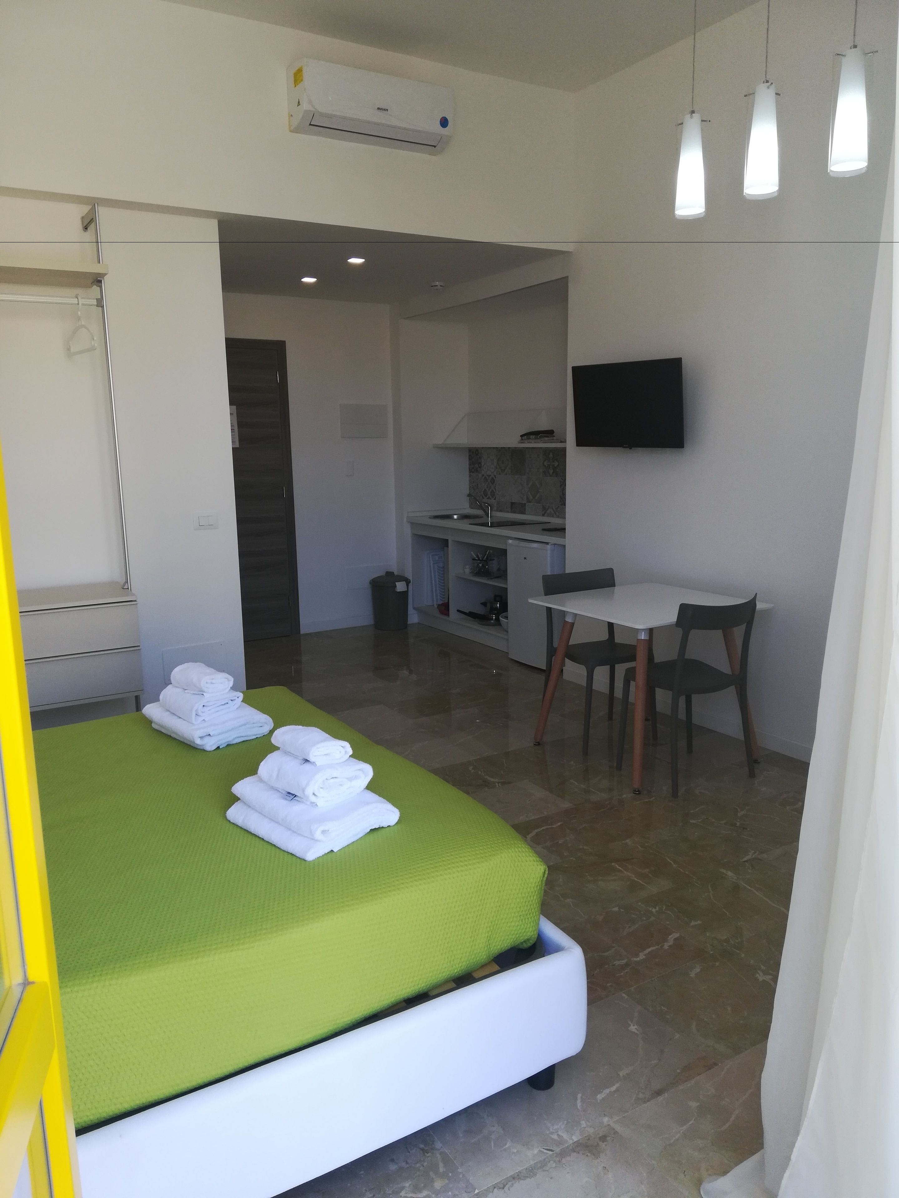 Habitación de hotel con una cama, un edredón verde, un escritorio con una silla, un televisor montado en la pared y una kitchenette.