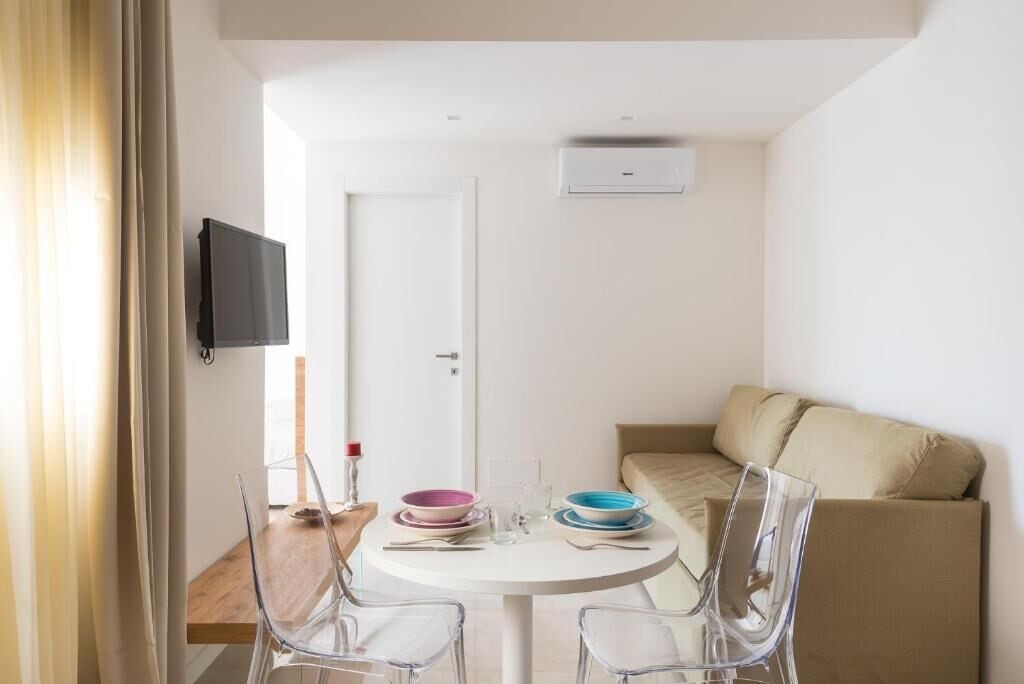 Un espacio habitable pequeño y moderno con una mesa de comedor redonda, sillas transparentes, un sofá y un televisor de pantalla plana montado en la pared.