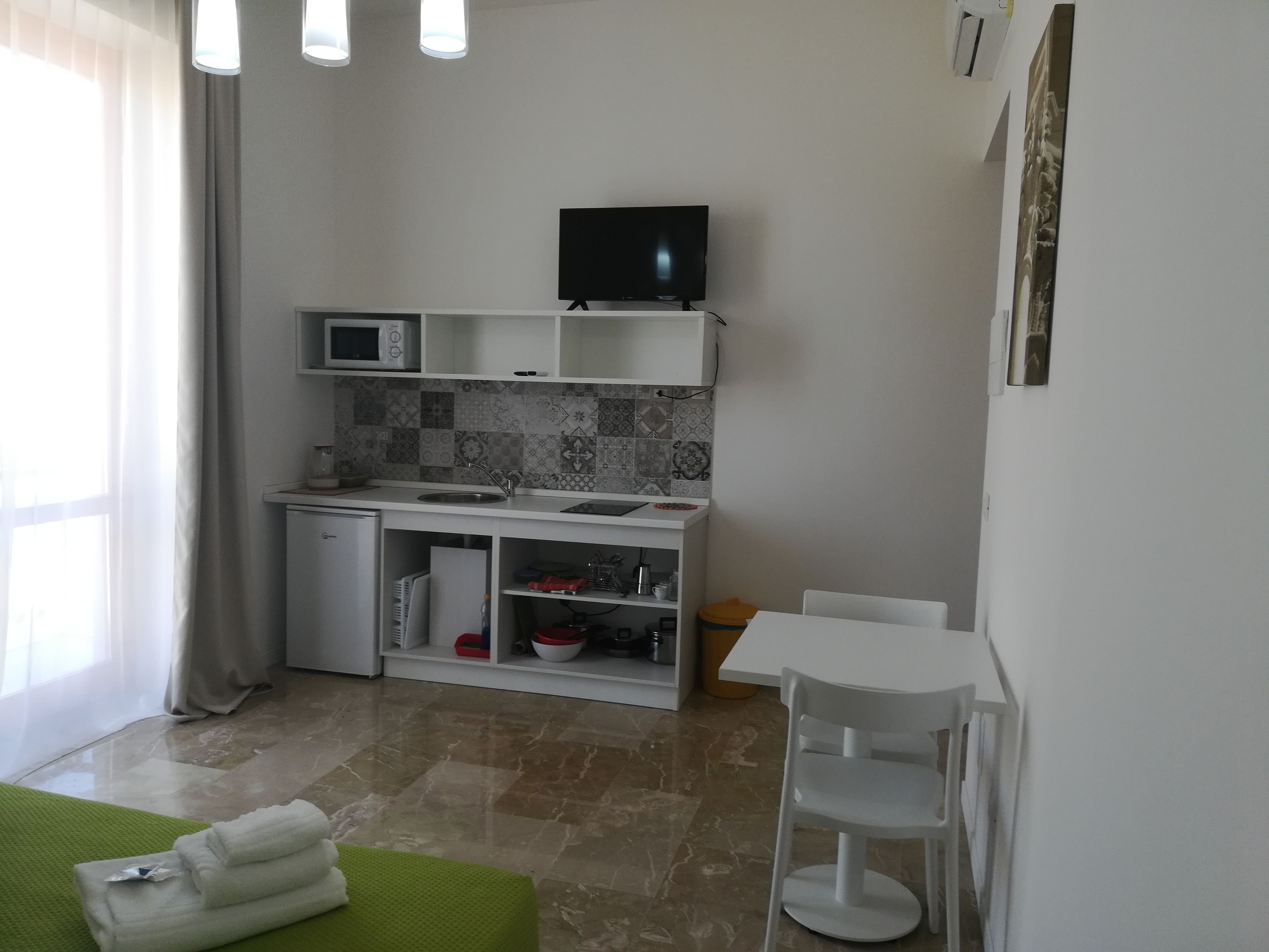 A compact kitchen with a microwave, sink, and a small refrigerator. There is a white table and a chair, and a television mounted above the counter.