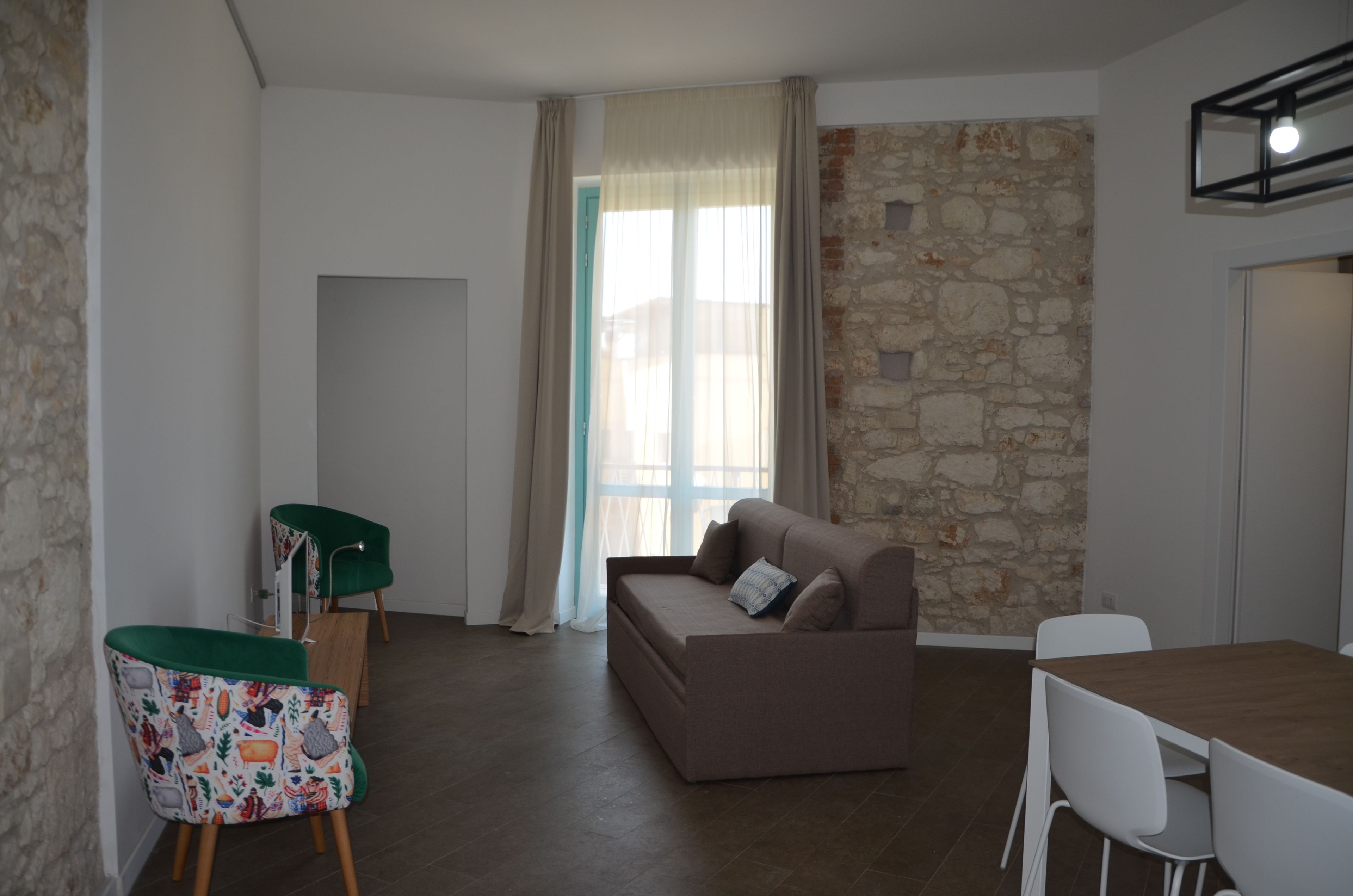 Una sala moderna con un sofá, una mesa de comedor y una silla estampada. Se ve una pared de piedra y una ventana con cortinas.