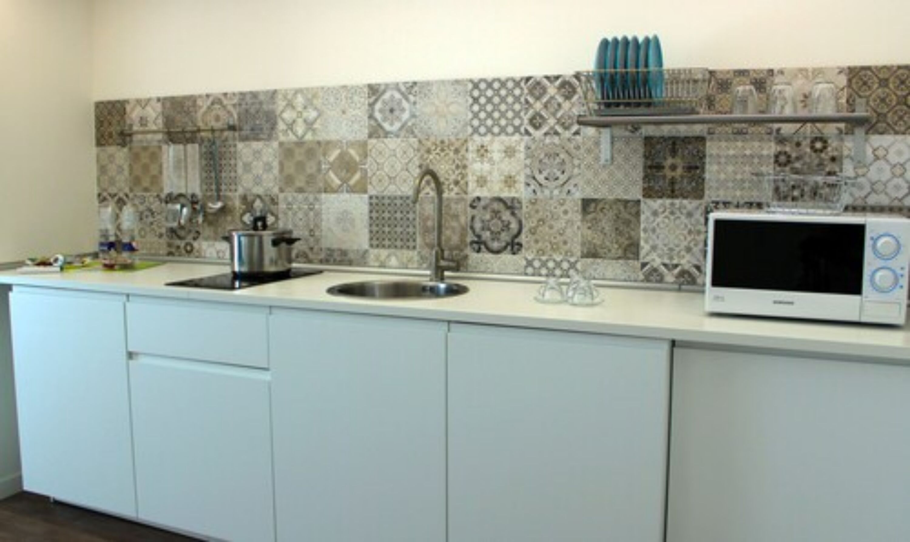 A kitchen with a tiled backsplash, a stainless steel sink, a microwave, and a dish rack.