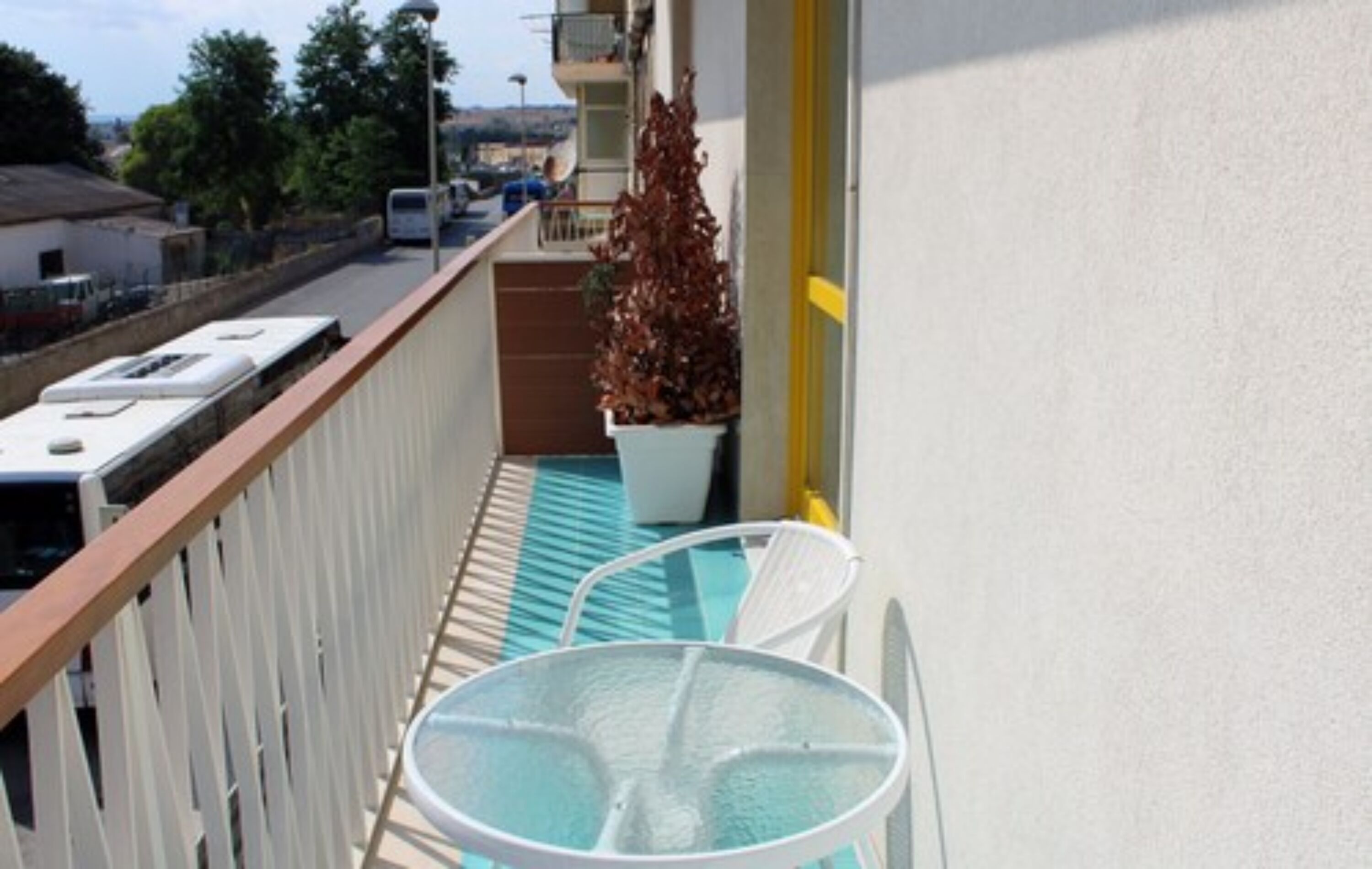A balcony with a table and chairs, a potted plant, and a view of the street and buildings in the background.