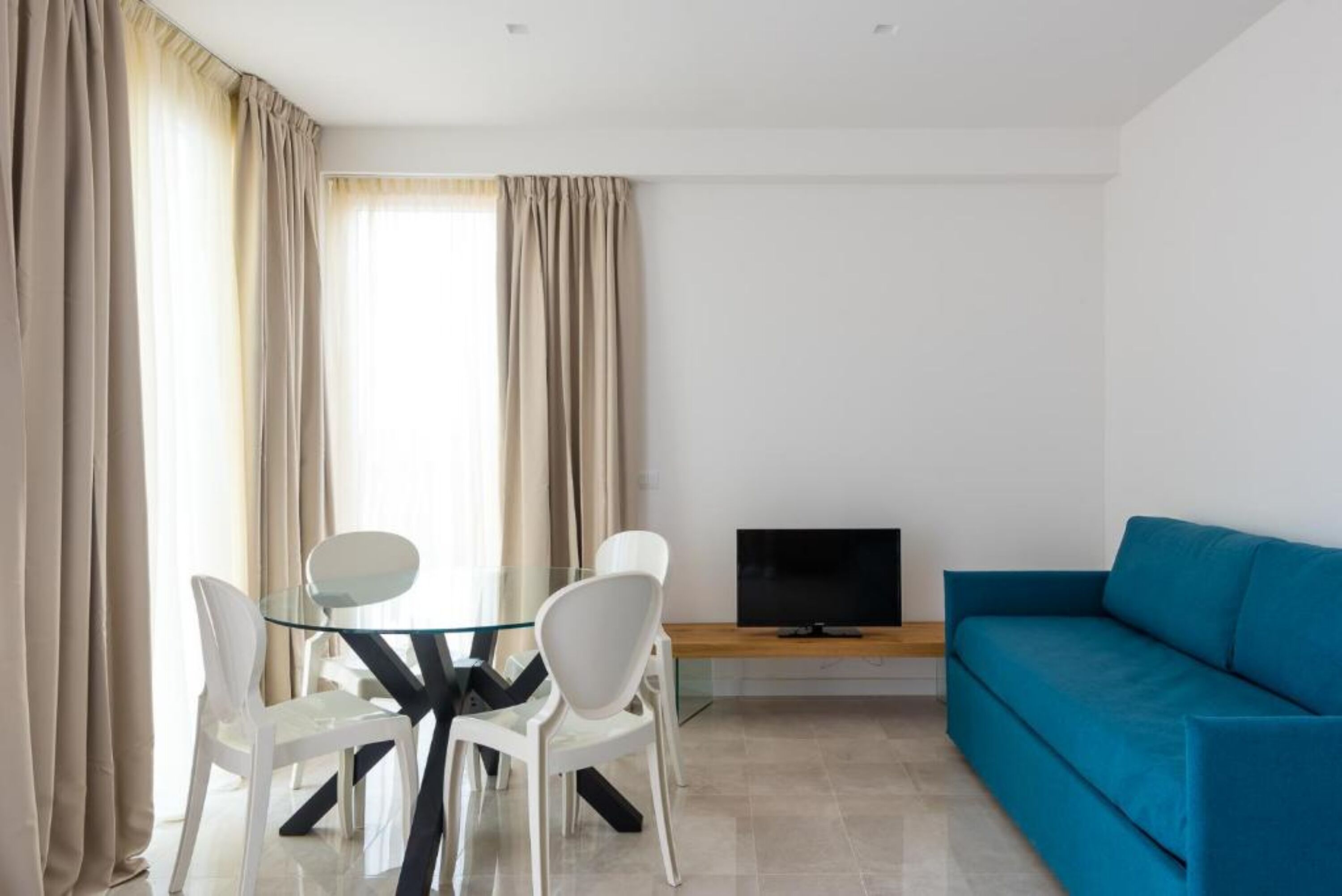 Uma sala de estar moderna com uma mesa de jantar de vidro, cadeiras brancas, um sofá azul e uma televisão de tela plana em um suporte de madeira.