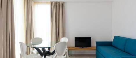 Uma sala de estar moderna com uma mesa de jantar de vidro, cadeiras brancas, um sofá azul e uma televisão de tela plana em um suporte de madeira.