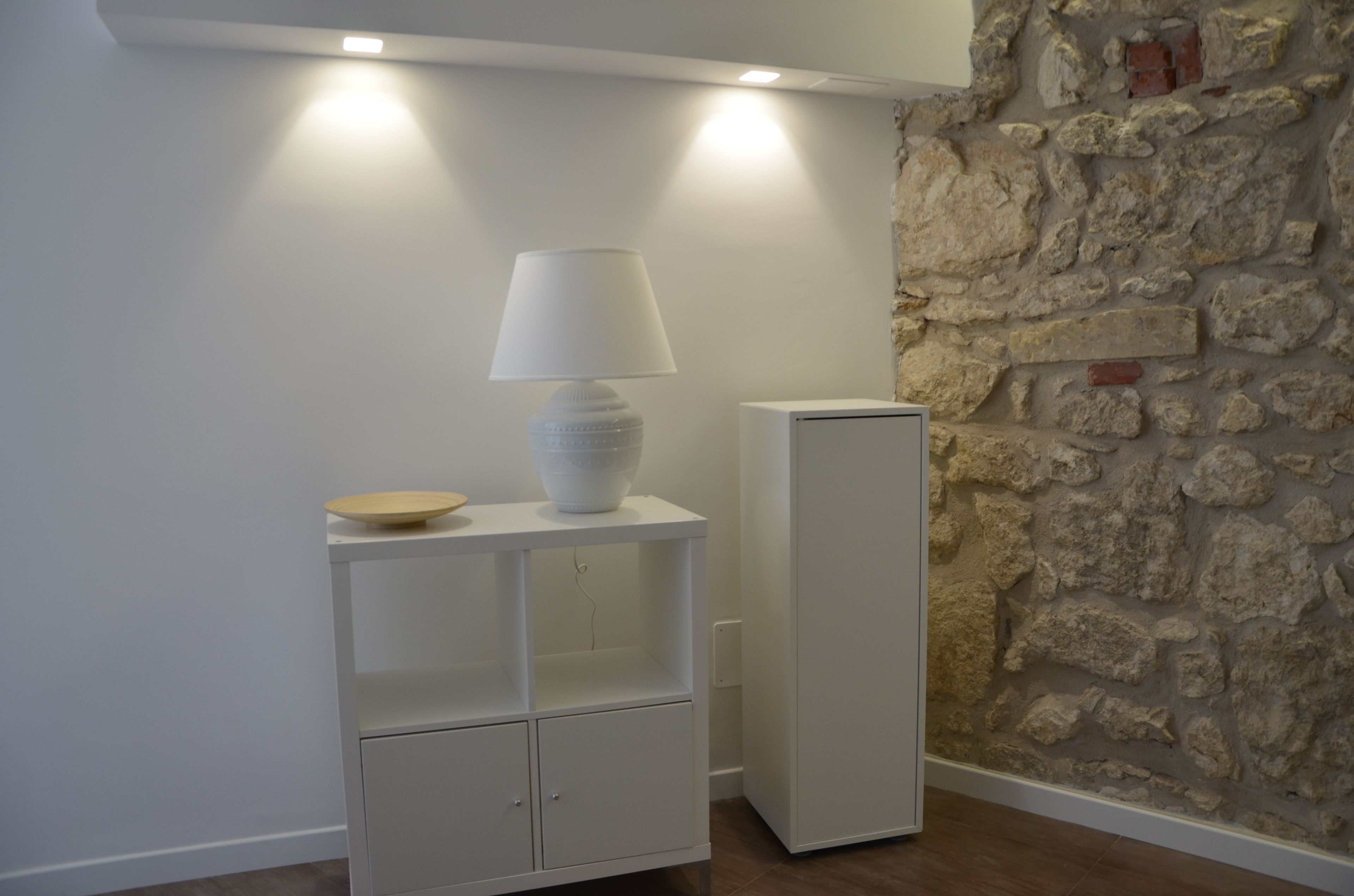 Un meuble blanc sur lequel sont posés une lampe et un bol, un réfrigérateur blanc et un mur en pierre.