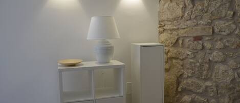 Un meuble blanc sur lequel sont posés une lampe et un bol, un réfrigérateur blanc et un mur en pierre.