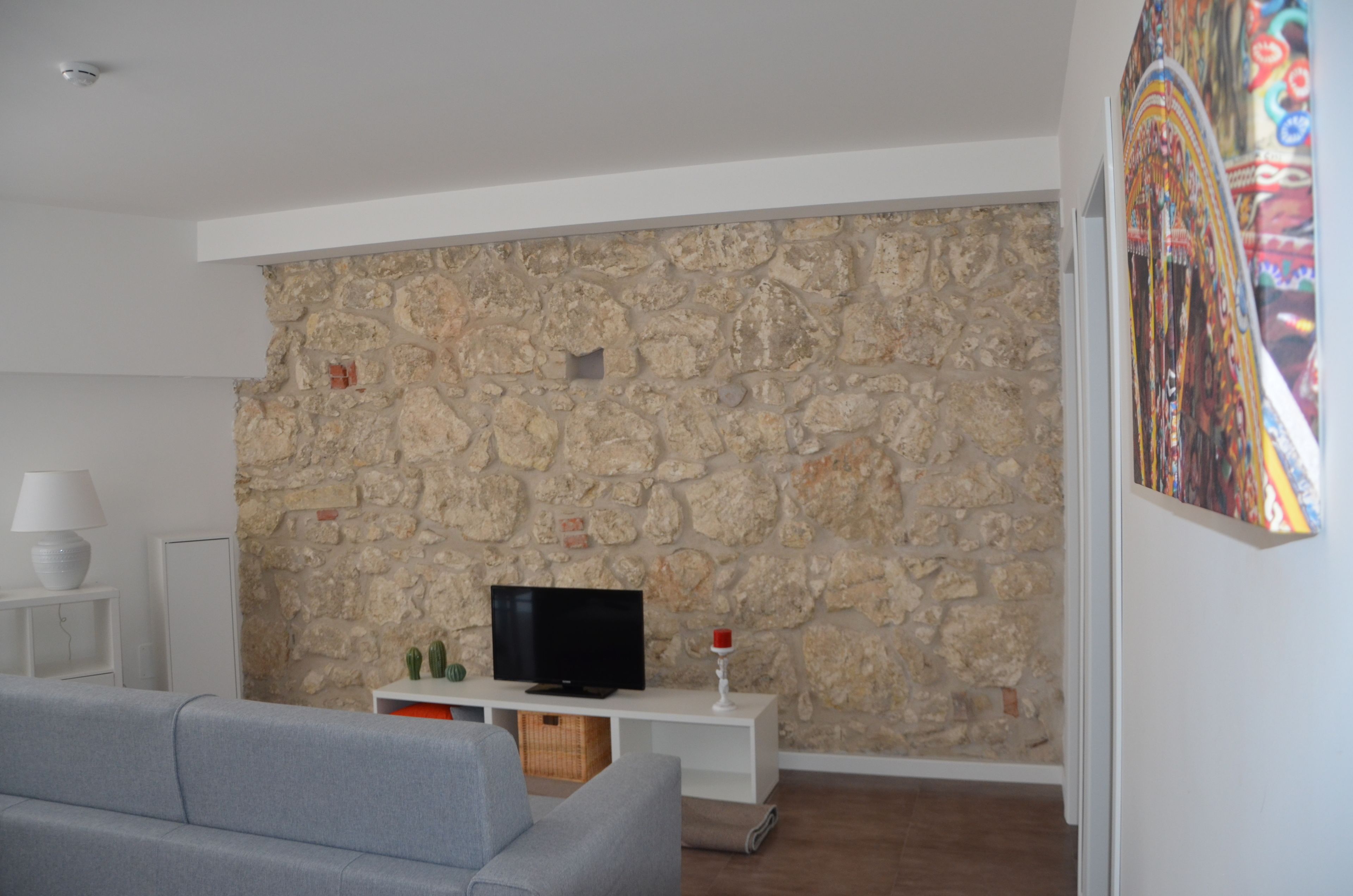 A living room with a stone wall, a flat-screen TV on a white stand, a grey sofa, and a small table with a lamp and decorative items.