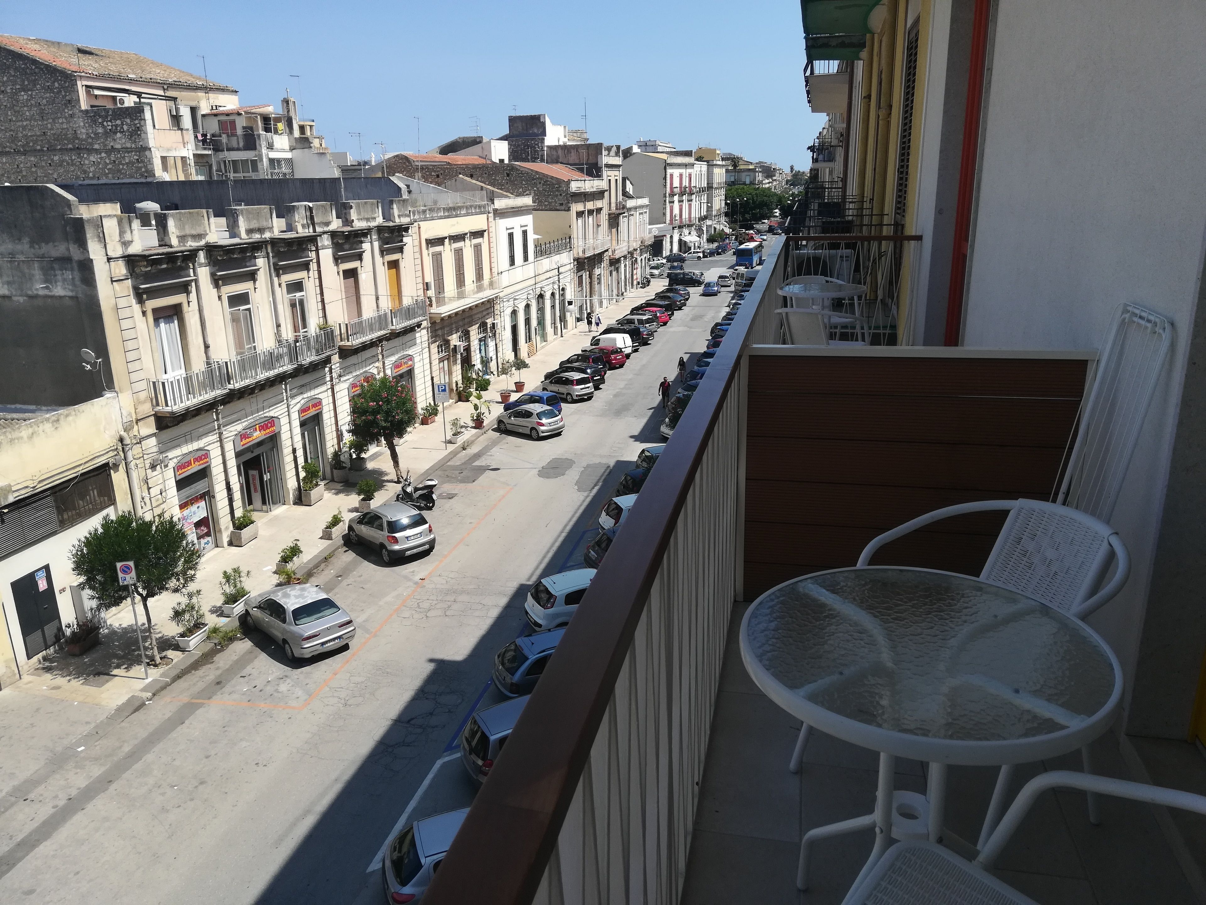 A balcony with white wicker chairs and a table, overlooking a street with parked cars and buildings.