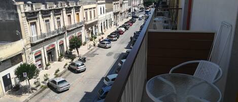A balcony with white wicker chairs and a table, overlooking a street with parked cars and buildings.