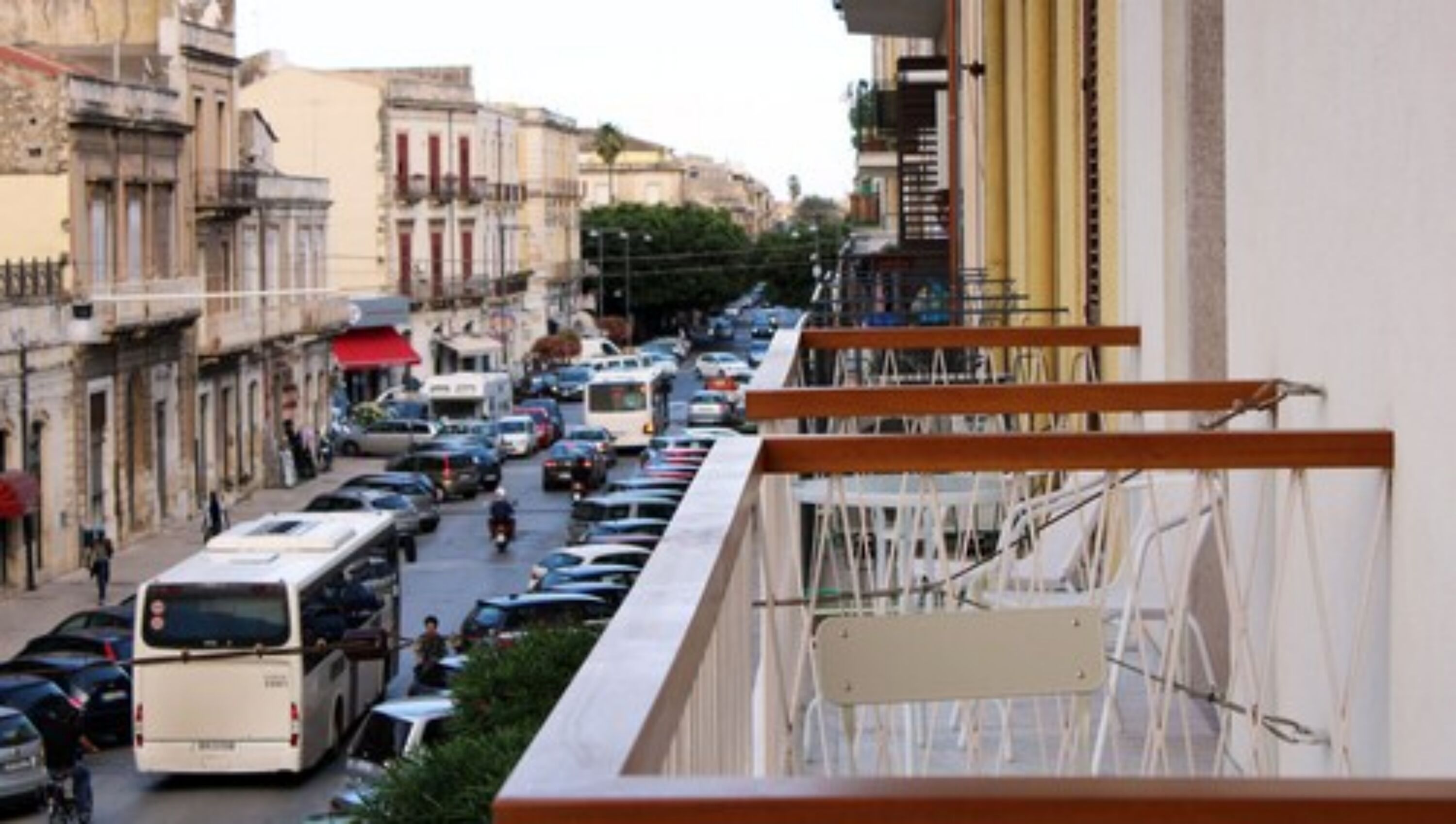 Vista de la calle con coches estacionados y un autobús, edificios con balcones y cielo despejado.