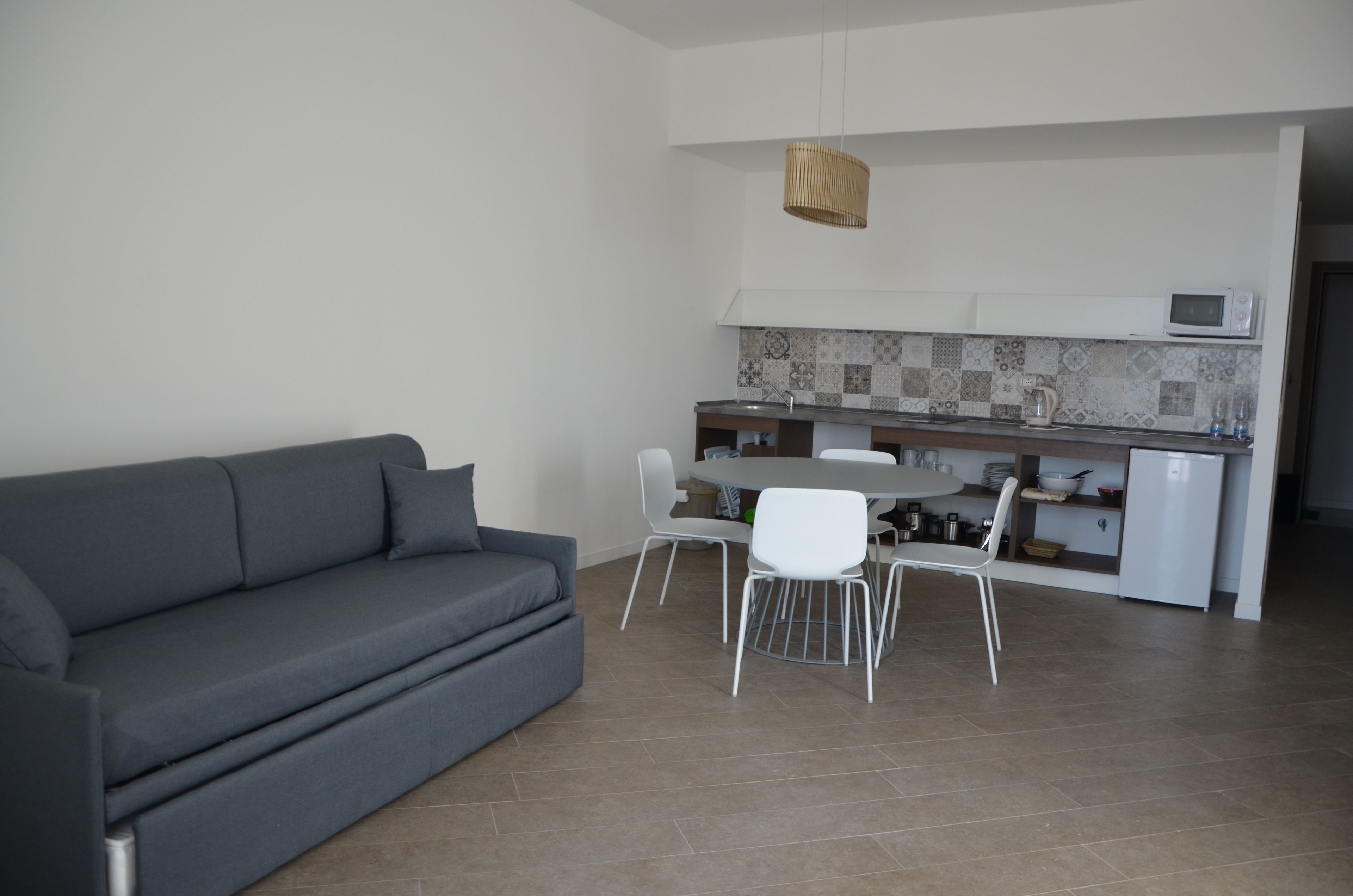 Un espacio habitable moderno con un sofá gris, una mesa de comedor con sillas, una zona de cocina con microondas y una lámpara colgante.