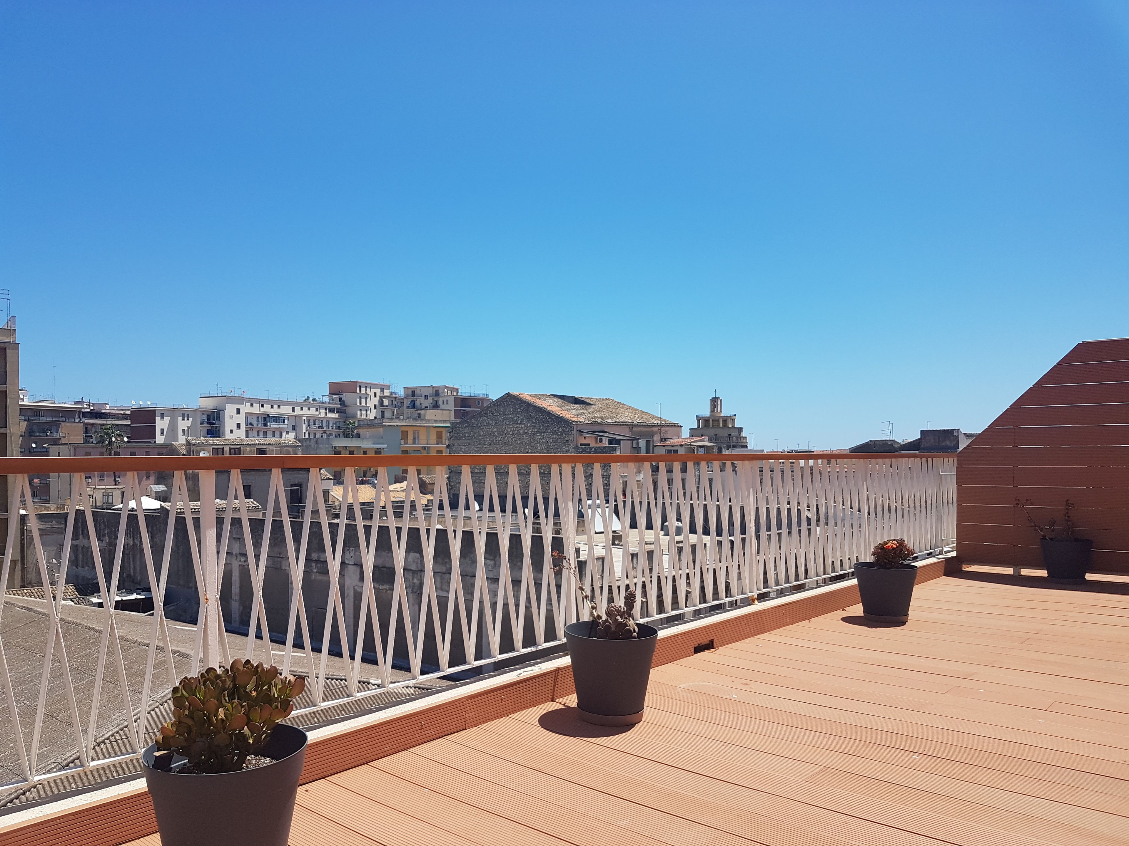 A rooftop terrace with a wooden floor, white railing, and potted plants.
