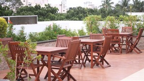 Eine Dachterrasse mit Holztischen und -stühlen, umgeben von Grünpflanzen und einer weißen Mauer.