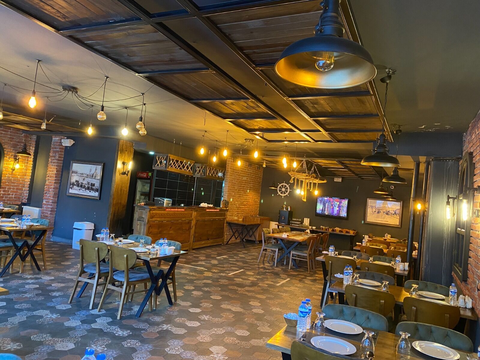 Un restaurante con suelo de patrón hexagonal, mesas y sillas de madera, lámparas colgantes y paredes de ladrillo.