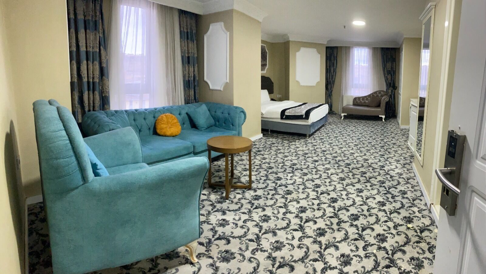 Quarto de hotel com sofá azul, uma mesinha redonda, cama com colchão branco e carpete estampado.