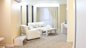A modern, well-lit living room with a white sofa, a coffee table, and a small white desk. The room features patterned carpet, a large window with curtains, and a wall-mounted air conditioning unit. - Buyuk Kale Hotel (Kars)