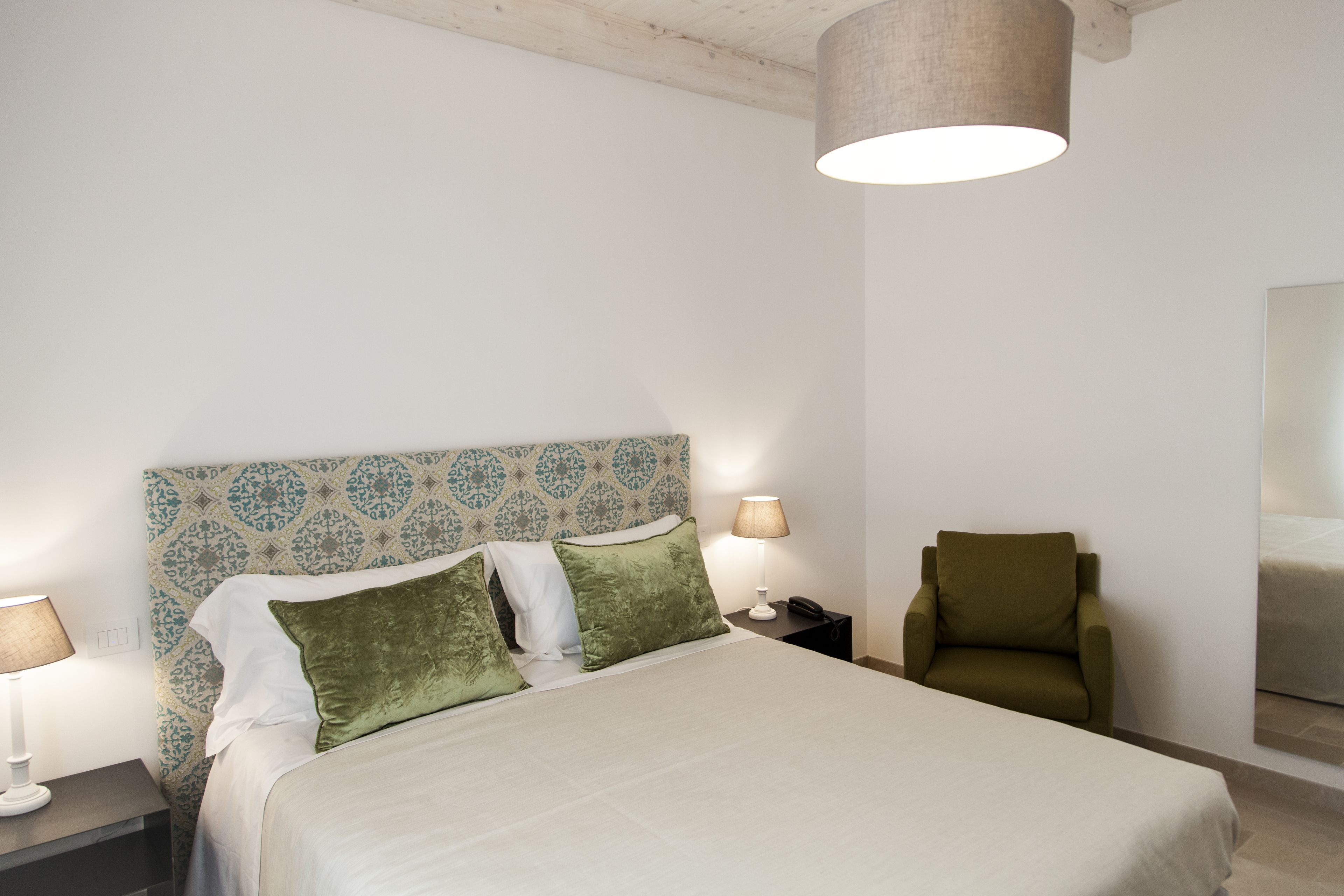 Un dormitorio ordenado con un cabecero estampado, una cama con almohadas verdes, lámparas de noche y una silla.