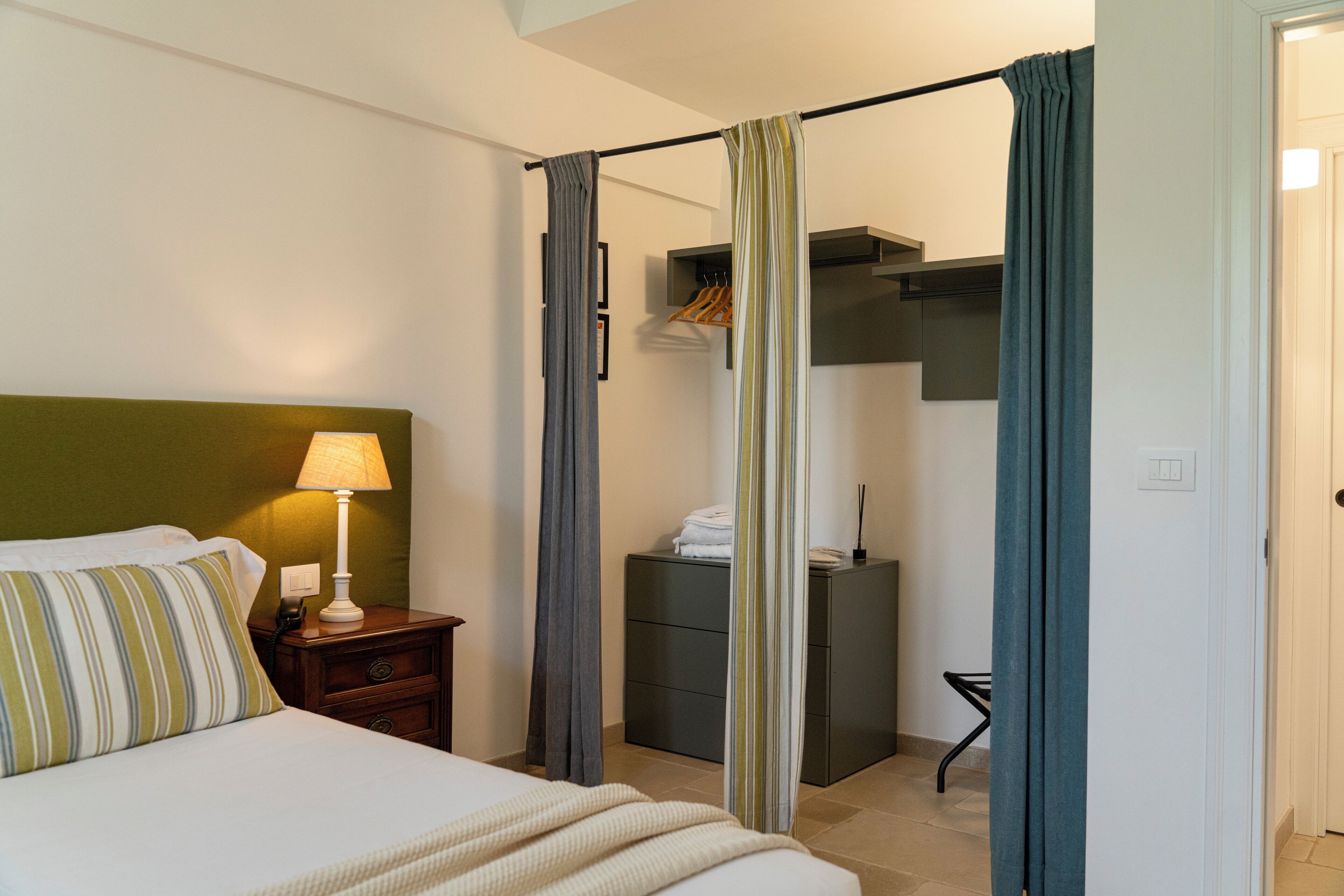 Quarto de hotel com cama, mesa de cabeceira com abajur, armário com roupas penduradas e banheiro visível através de uma cortina.