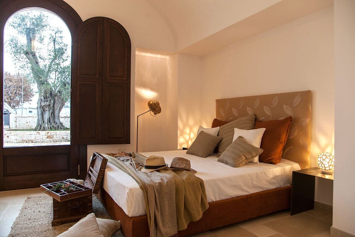 A bedroom with a large bed, a wooden headboard, a bedside table with a lamp, and a view of trees through a large arched window.