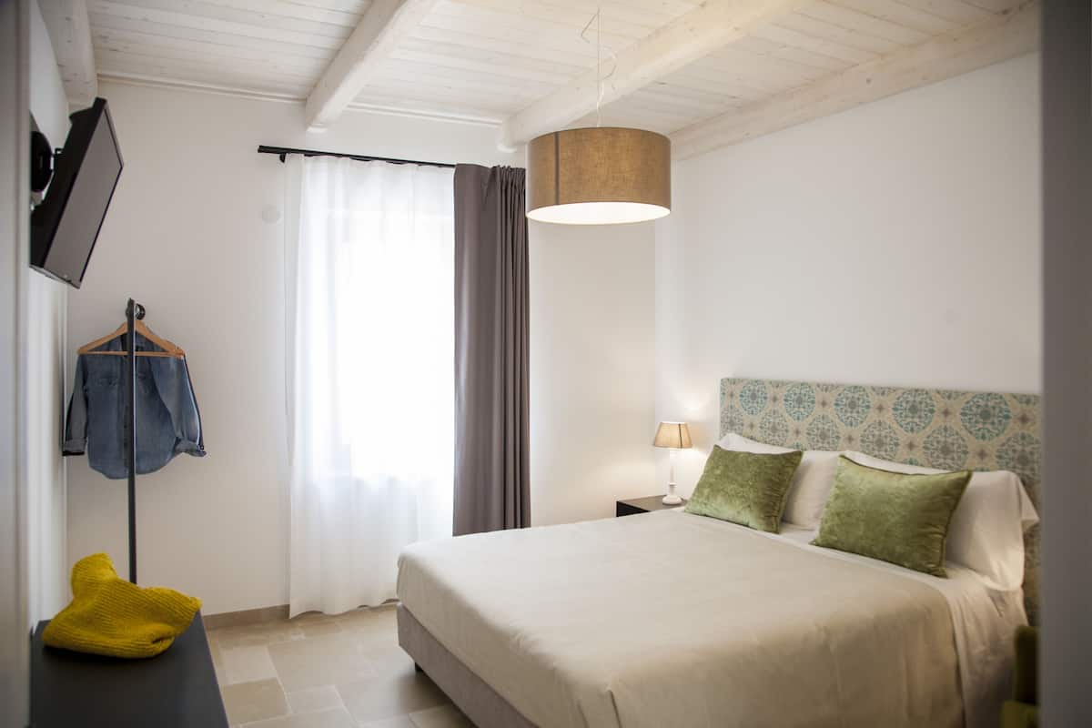 A bedroom with a bed, a hanging light fixture, a bedside table with a lamp, a coat rack with a jacket, and a window with curtains.