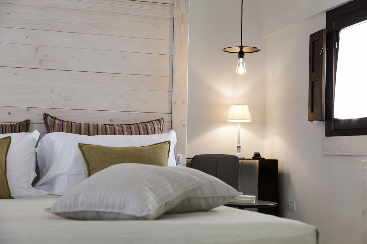 A hotel room with a wooden headboard, a bed with white bedding and green pillows, a bedside table with a lamp, and a chair.