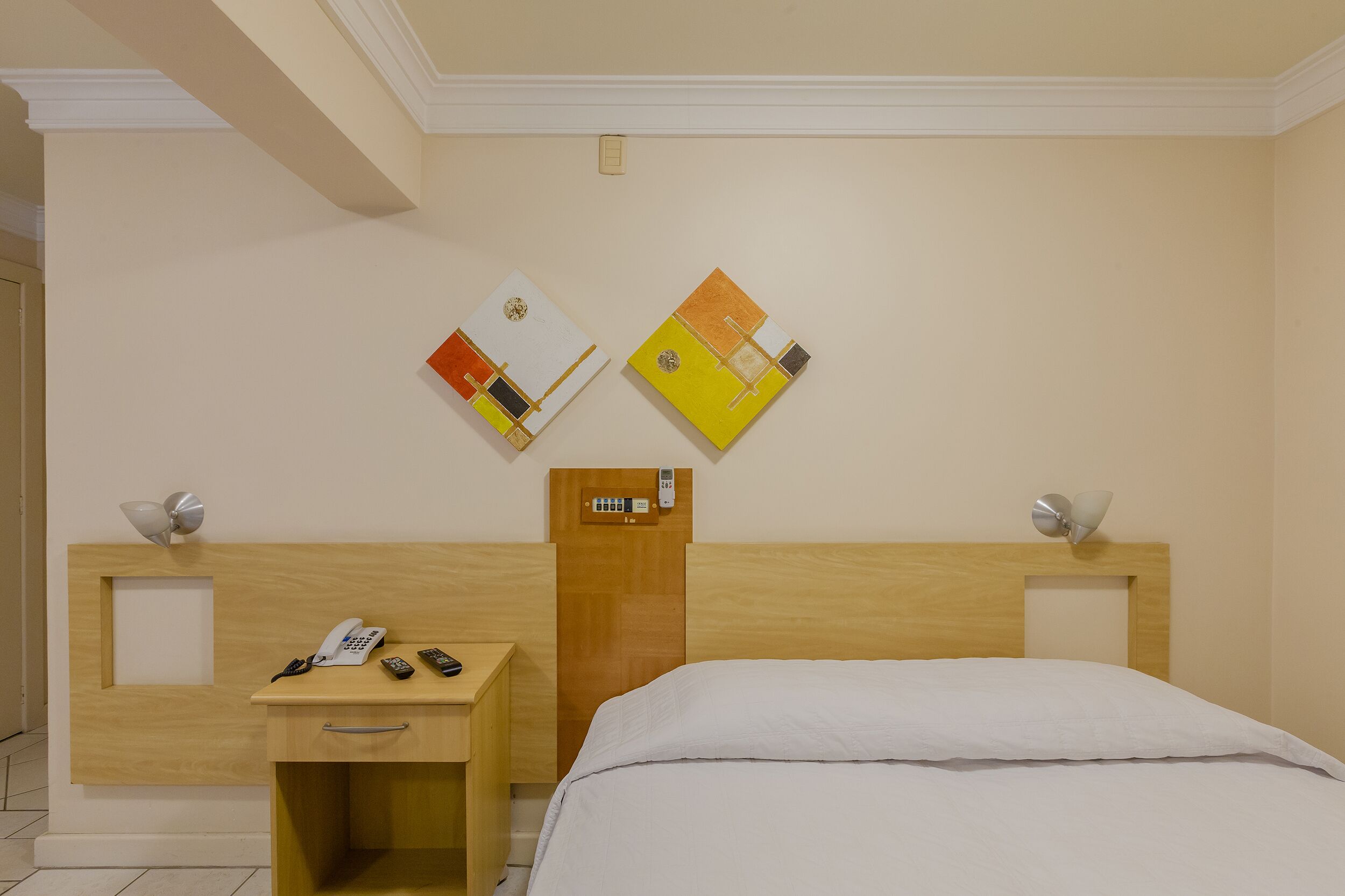 Habitación de hotel con cabecera de madera, mesita de noche con teléfono y controles remotos, y dos obras de arte colgadas en la pared.