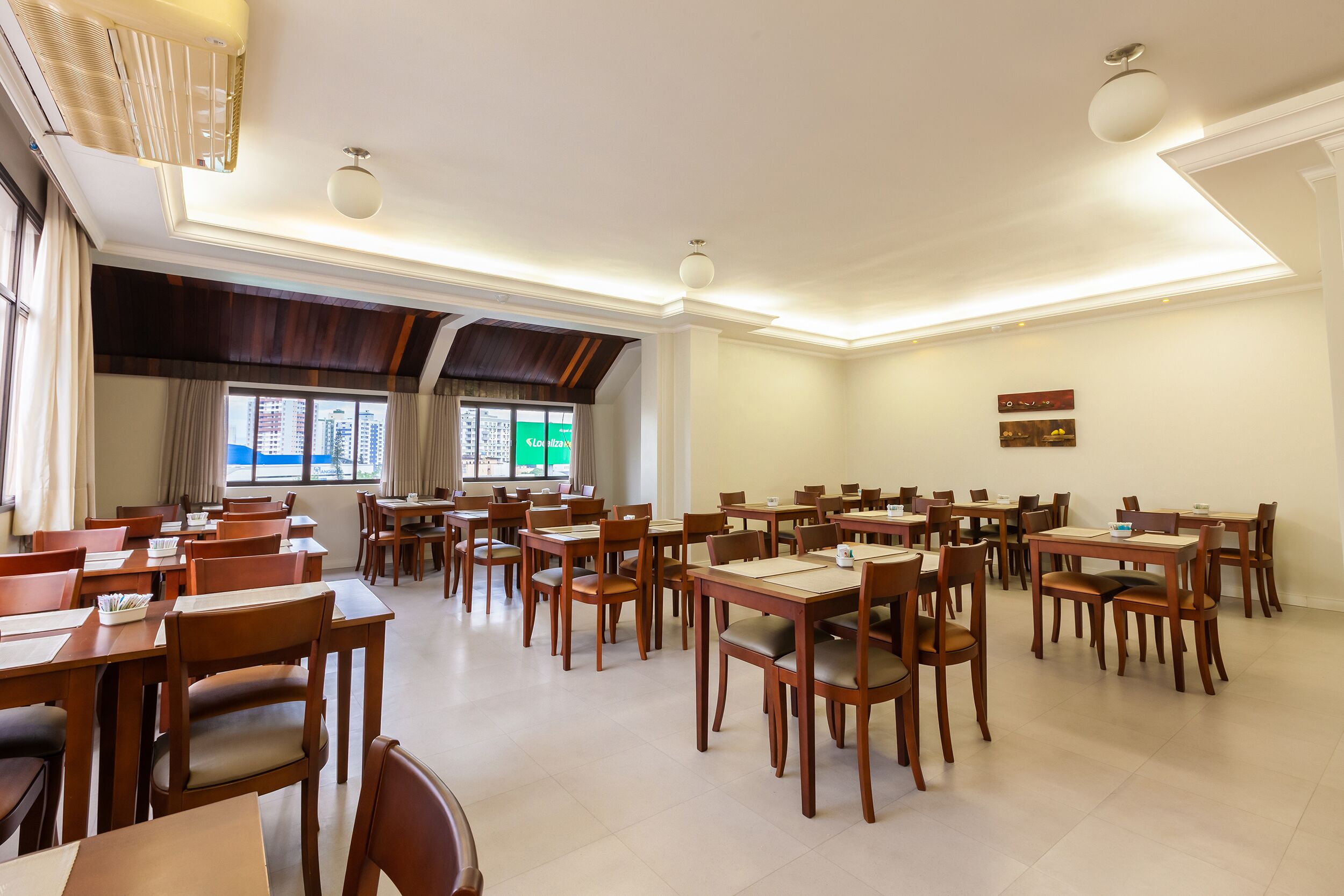 Un comedor amplio con varias mesas y sillas de madera, ventanas grandes con vistas a edificios e iluminación empotrada en el techo.