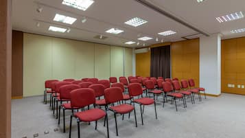Un salón de conferencias con hileras de sillas rojas, luces de techo y una gran pantalla de proyección.