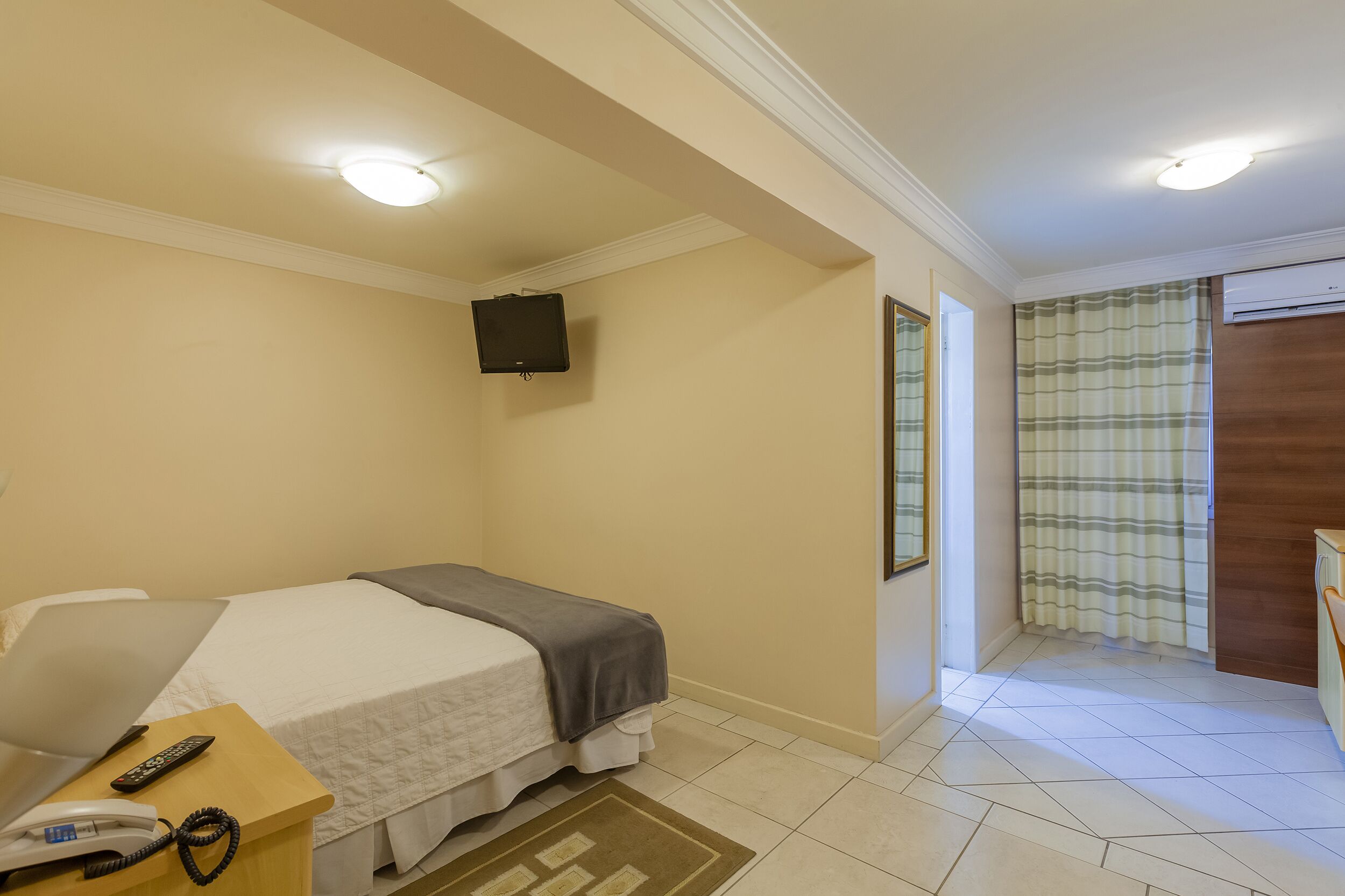 Une chambre d’hôtel comprenant un lit, un bureau avec un téléphone et une télécommande, un téléviseur fixé au mur et une salle de bain avec un rideau de douche.
