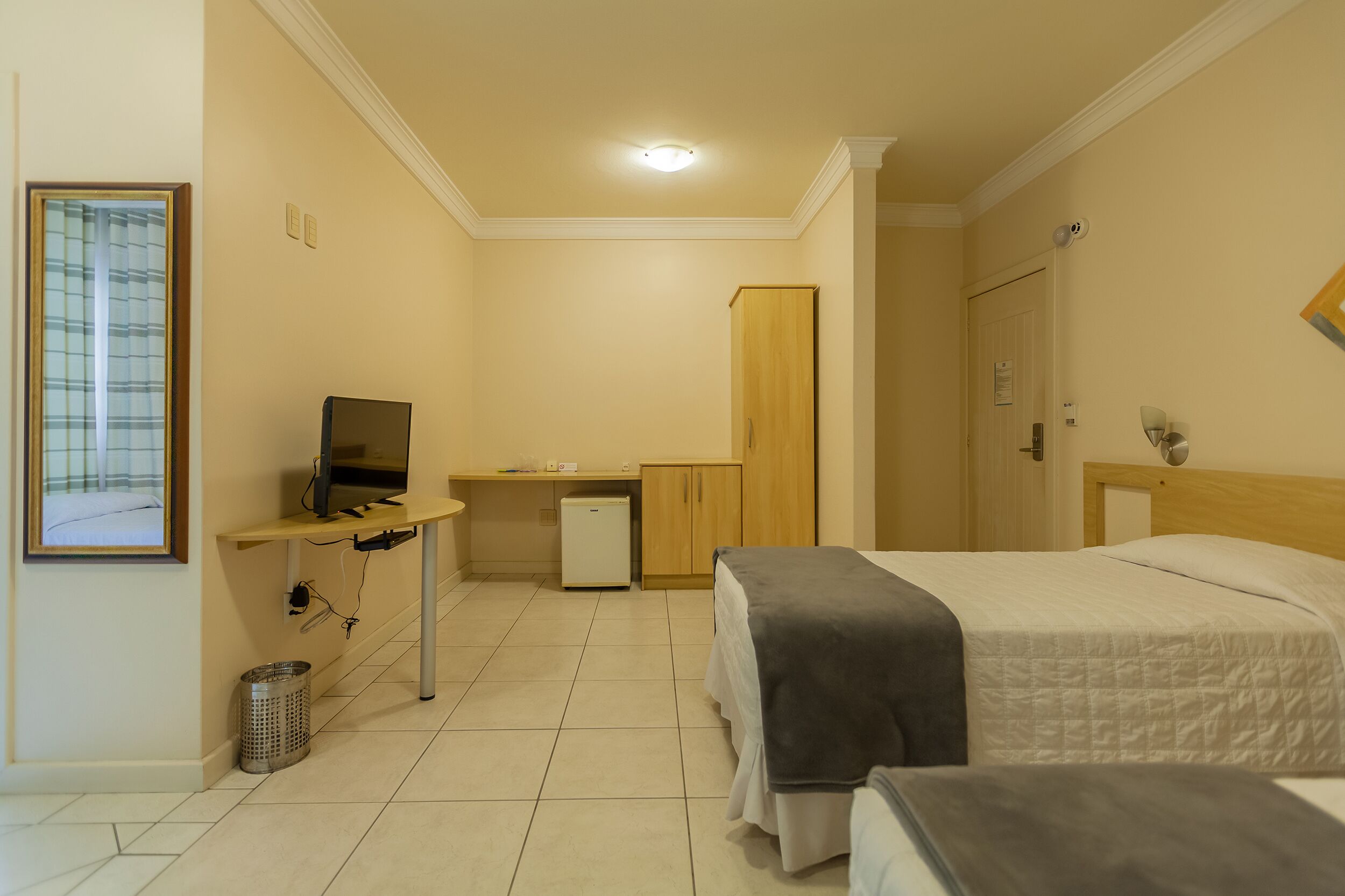 Habitación de hotel con cama, escritorio con computadora, refrigerador pequeño y ventana con persianas.