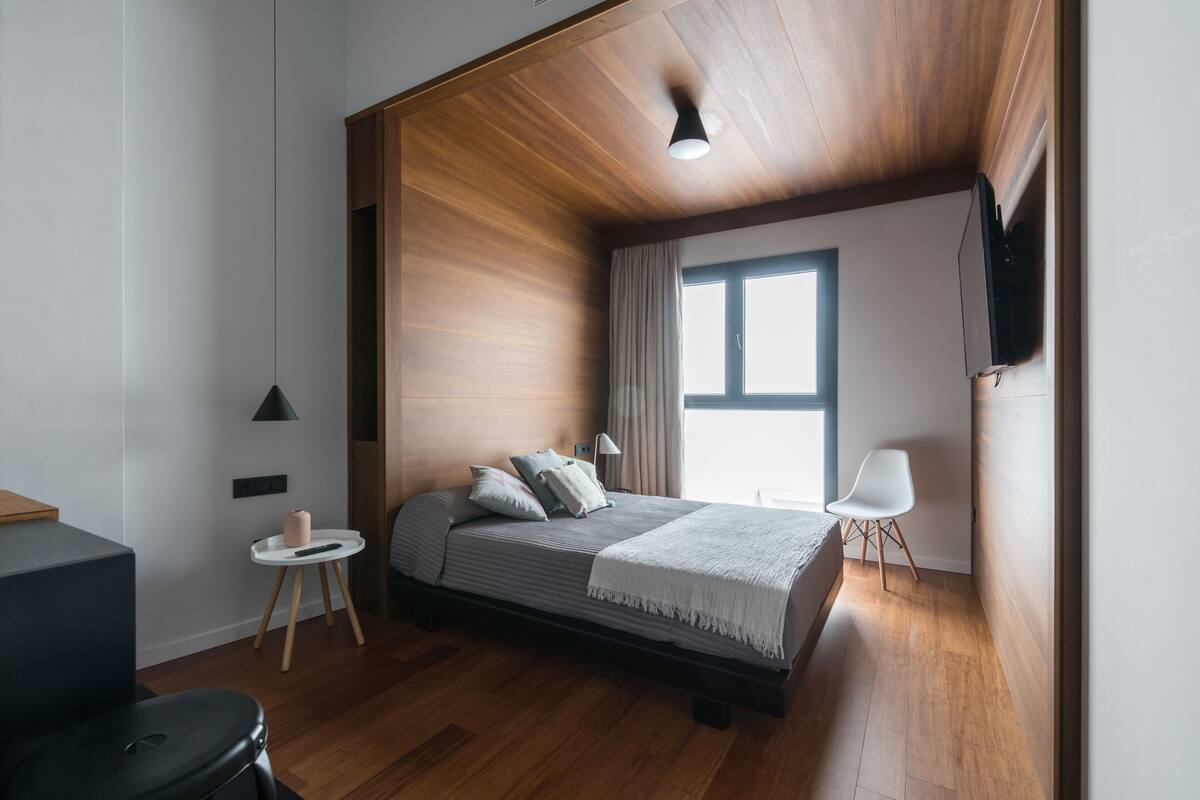 A modern bedroom with a wooden ceiling, a bed with pillows, a bedside table, a chair, and a window.